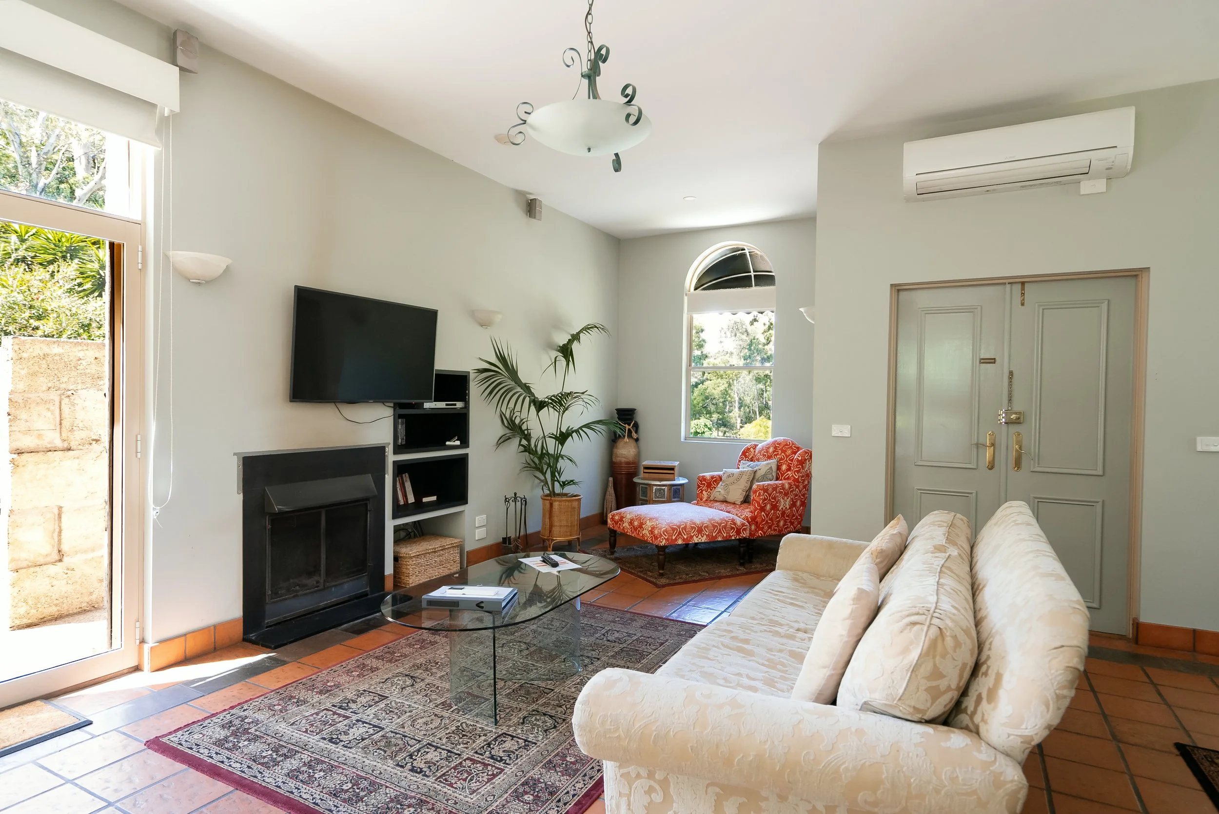 Living room with beige sofa, red armchair near window, wall-mounted TV, fireplace, black bookshelf, glass coffee table, patterned rug, large green plant, decorative vases, wooden floors, and closed front door.