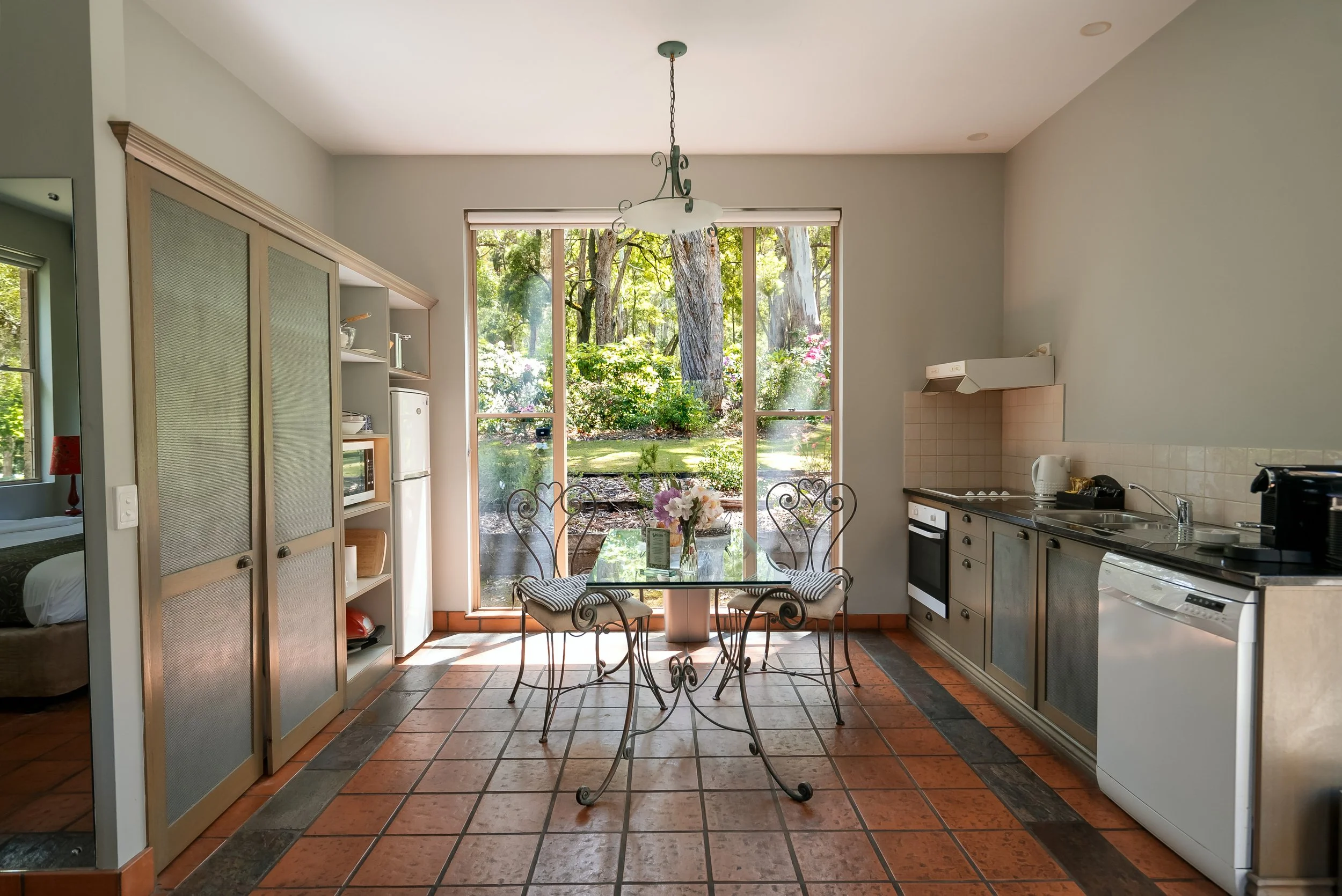 Kitchen with a glass dining table, metal chairs, large window overlooking a garden, kitchen appliances, and terracotta tiled floor.