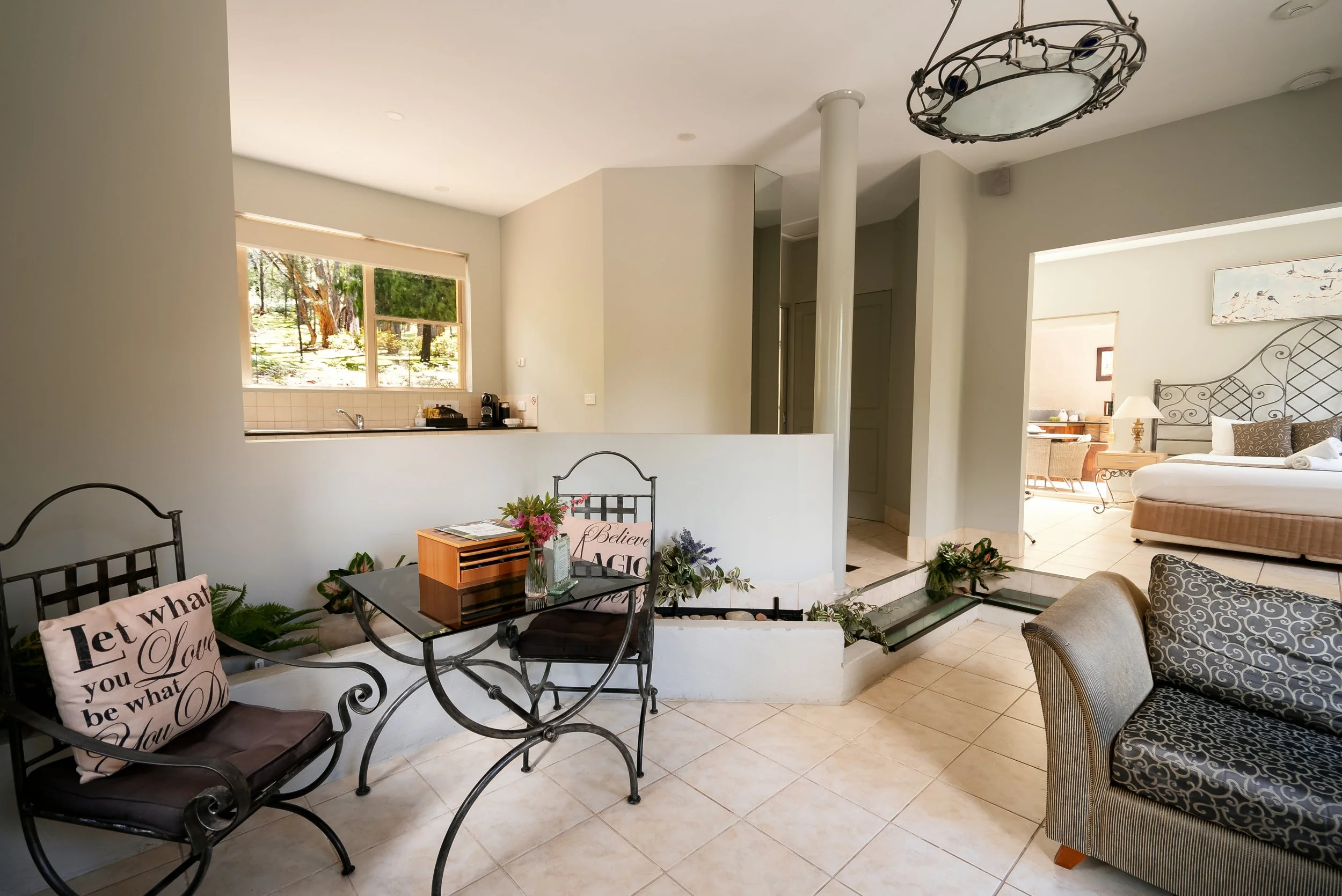 Living area with black wrought-iron chairs and a glass table with a cushion, a bedroom with a wrought-iron bedframe, and a hallway leading to a kitchen with a large window overlooking greenery.