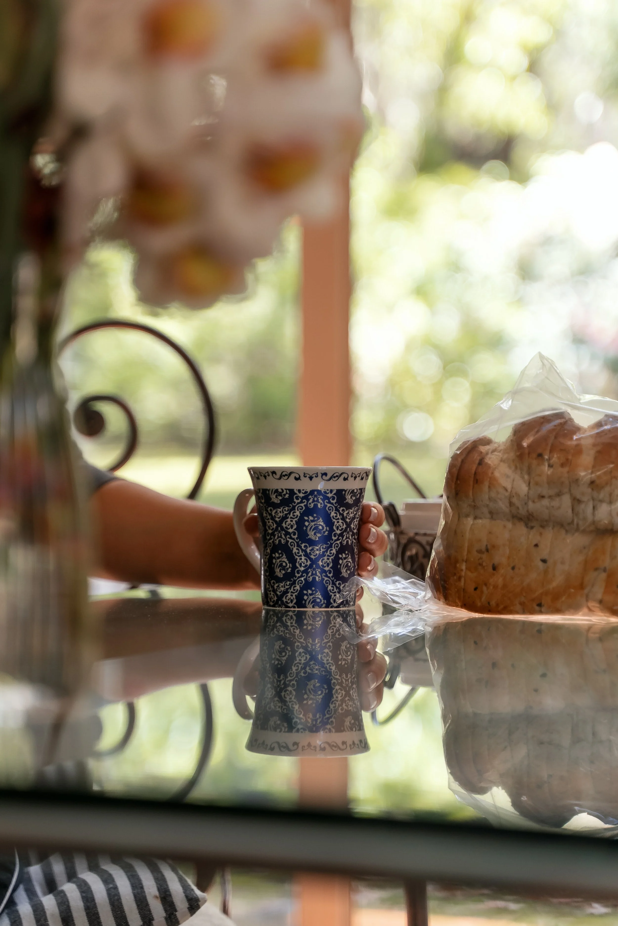 A person holding a blue and white patterned mug on a reflective glass table, with a large bag of bread rolls and blurred flowers and greenery in the background.