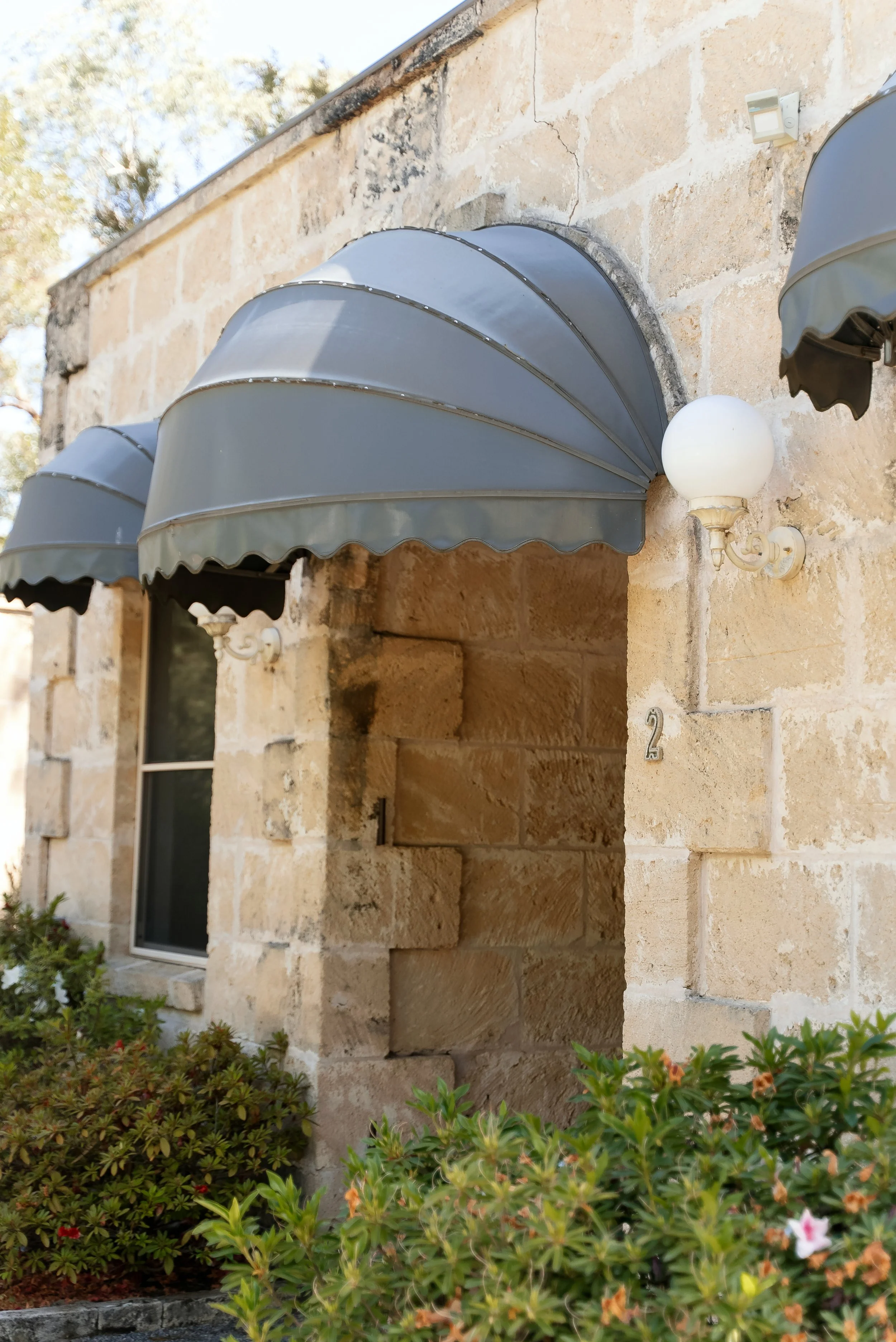 Close-up of a house exterior with two gray metal awnings over windows and an open doorway, and a white round wall light fixture with a scroll arm.
