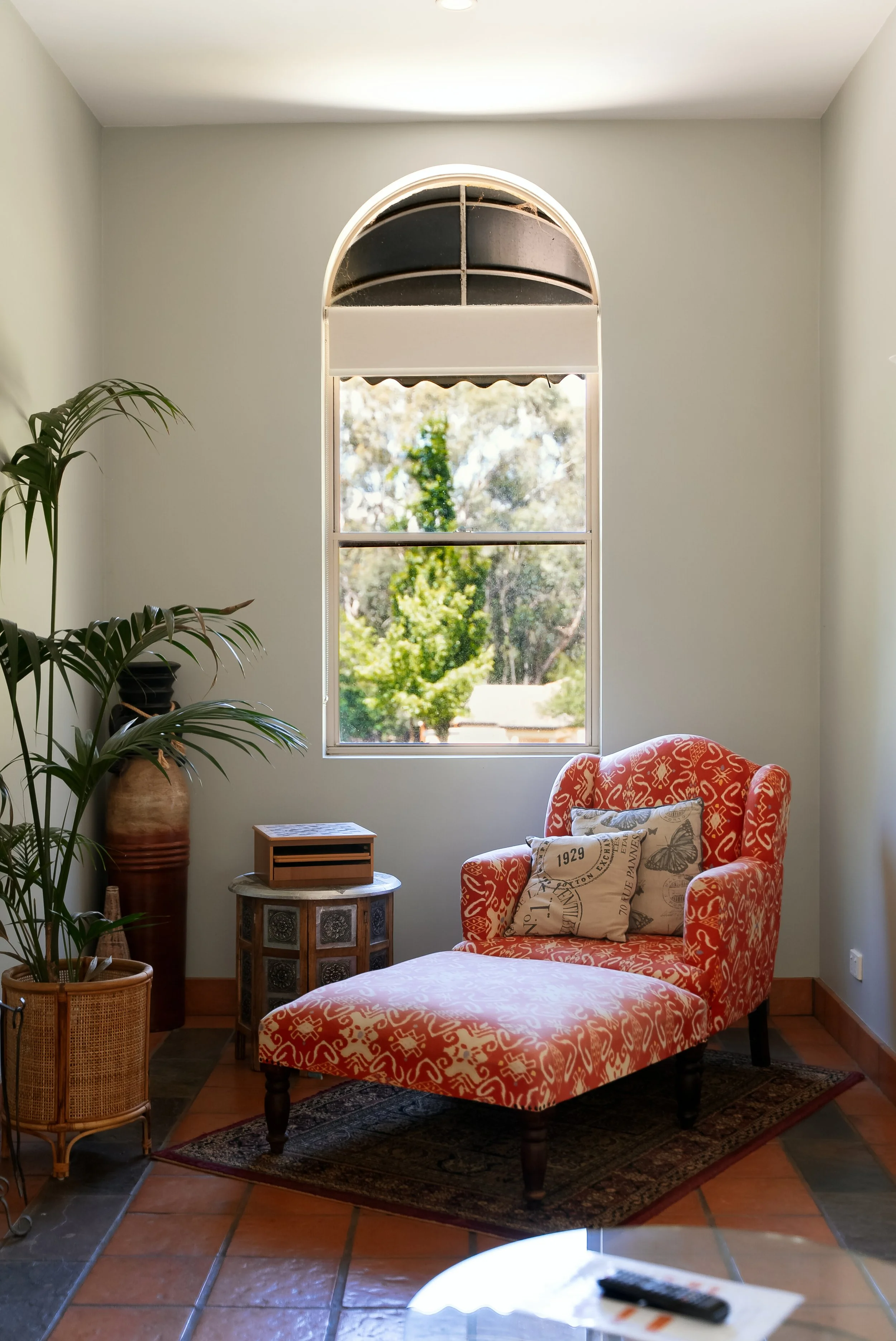 A cozy corner with a red patterned armchair and matching ottoman, a small side table with a decorative box, a large vase with a plant, and a window showing trees outside.