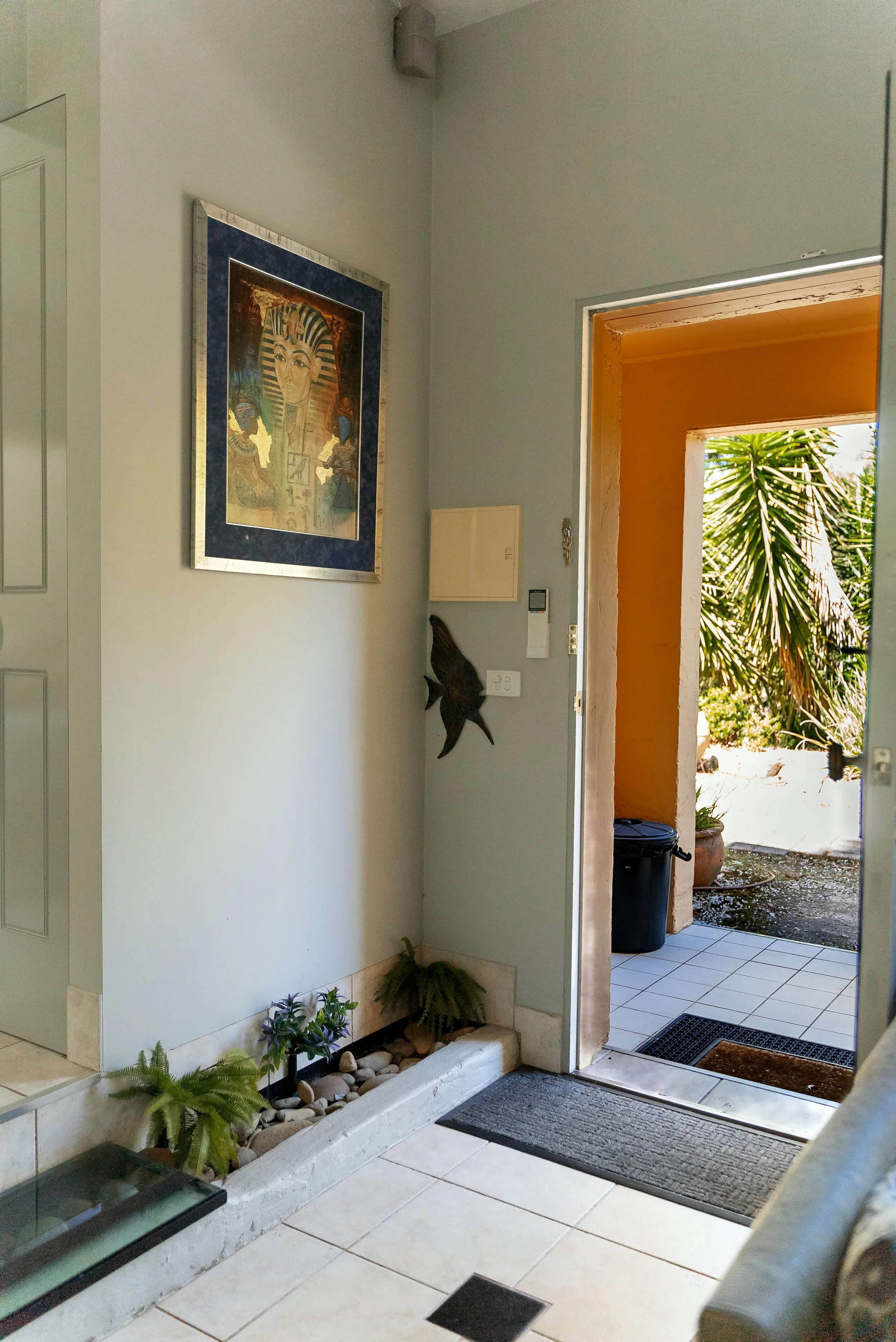 Interior view of a home entryway with a doorway leading outside, decorated with plants, framed artwork, and a wall-mounted fish sculpture.