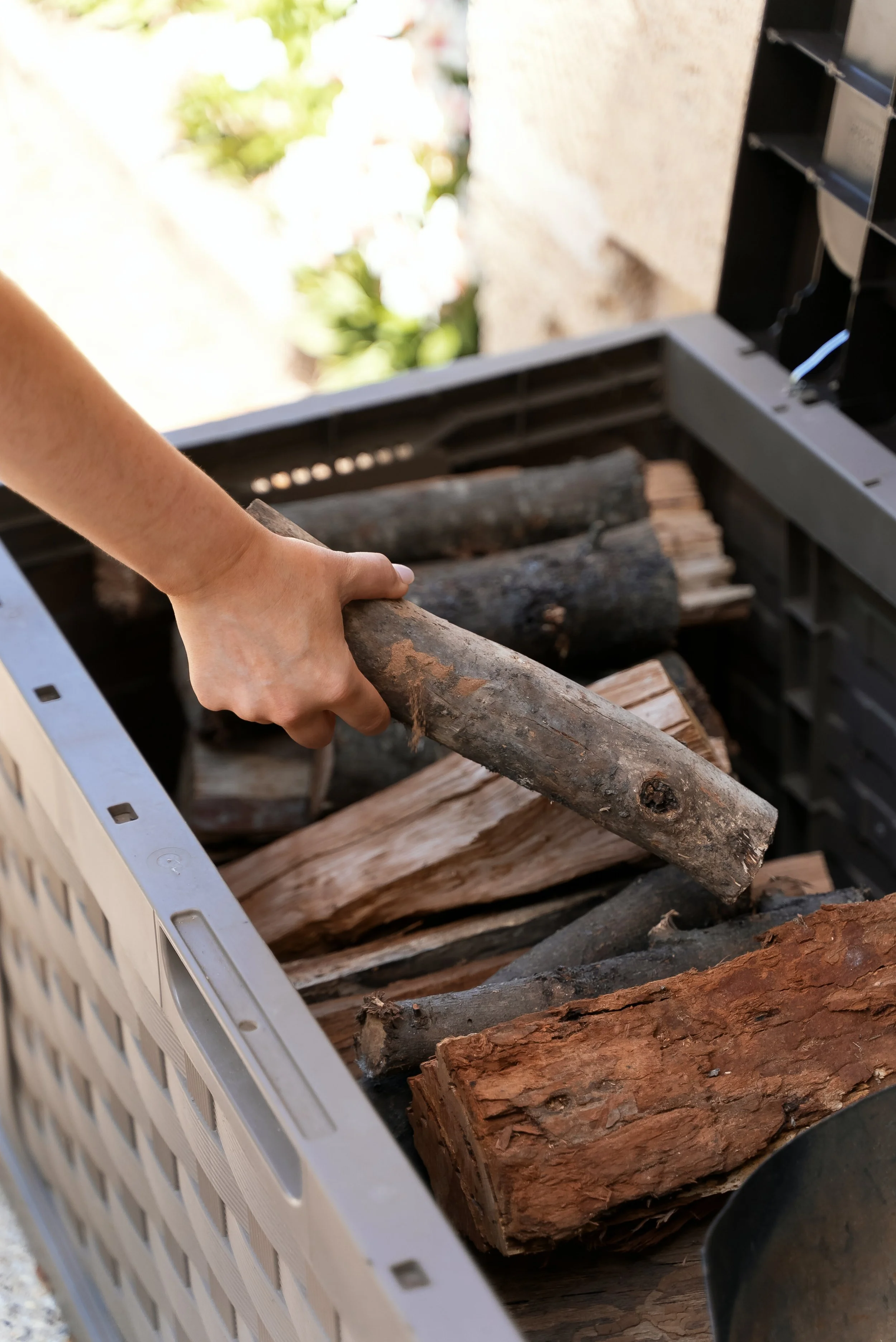A person is placing a piece of firewood into a wood storage container filled with various logs.