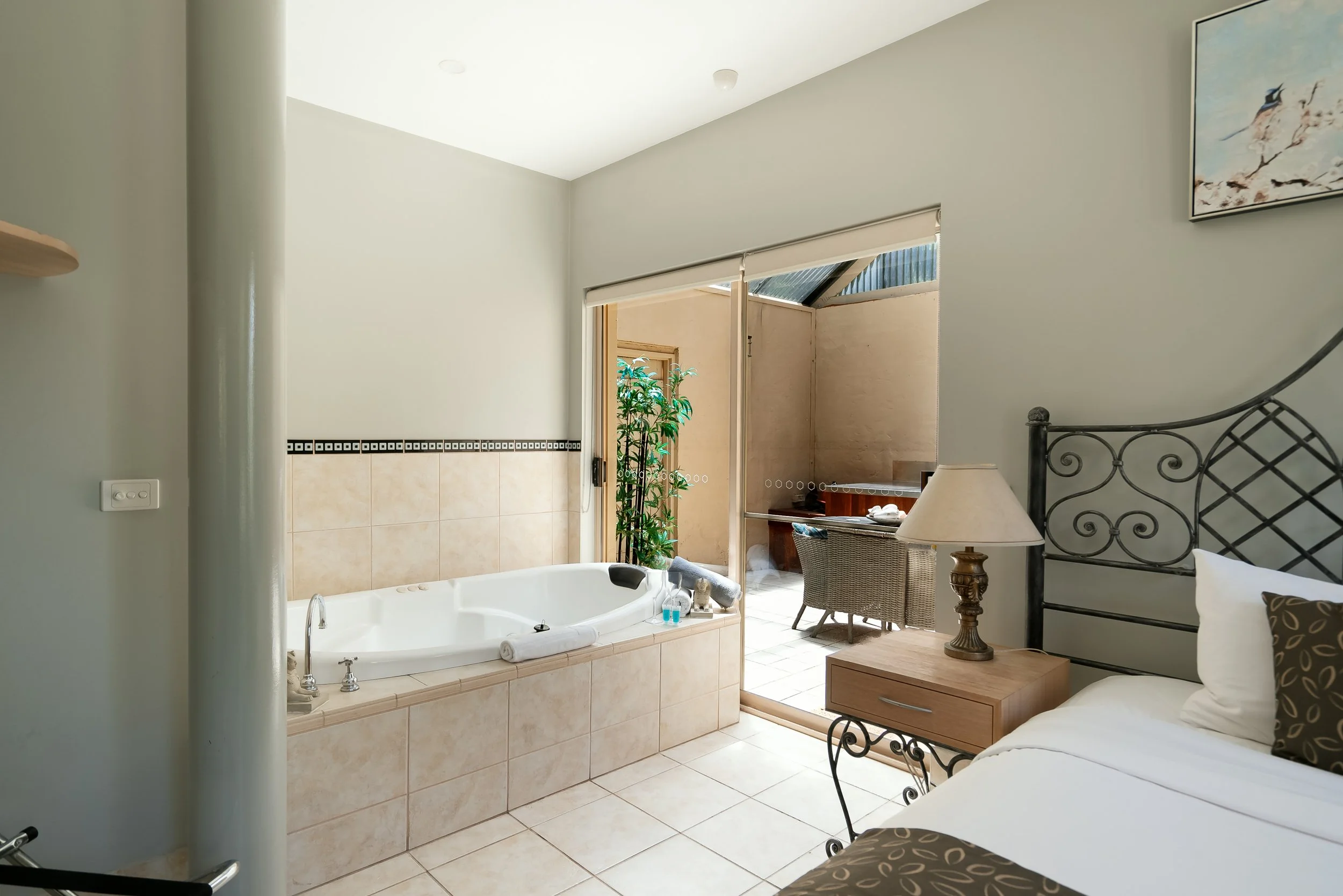 Hotel room with a large bathtub near a balcony door, a bed with decorative pillows, a nightstand with a lamp, and an outdoor sitting area with chairs and a small table.