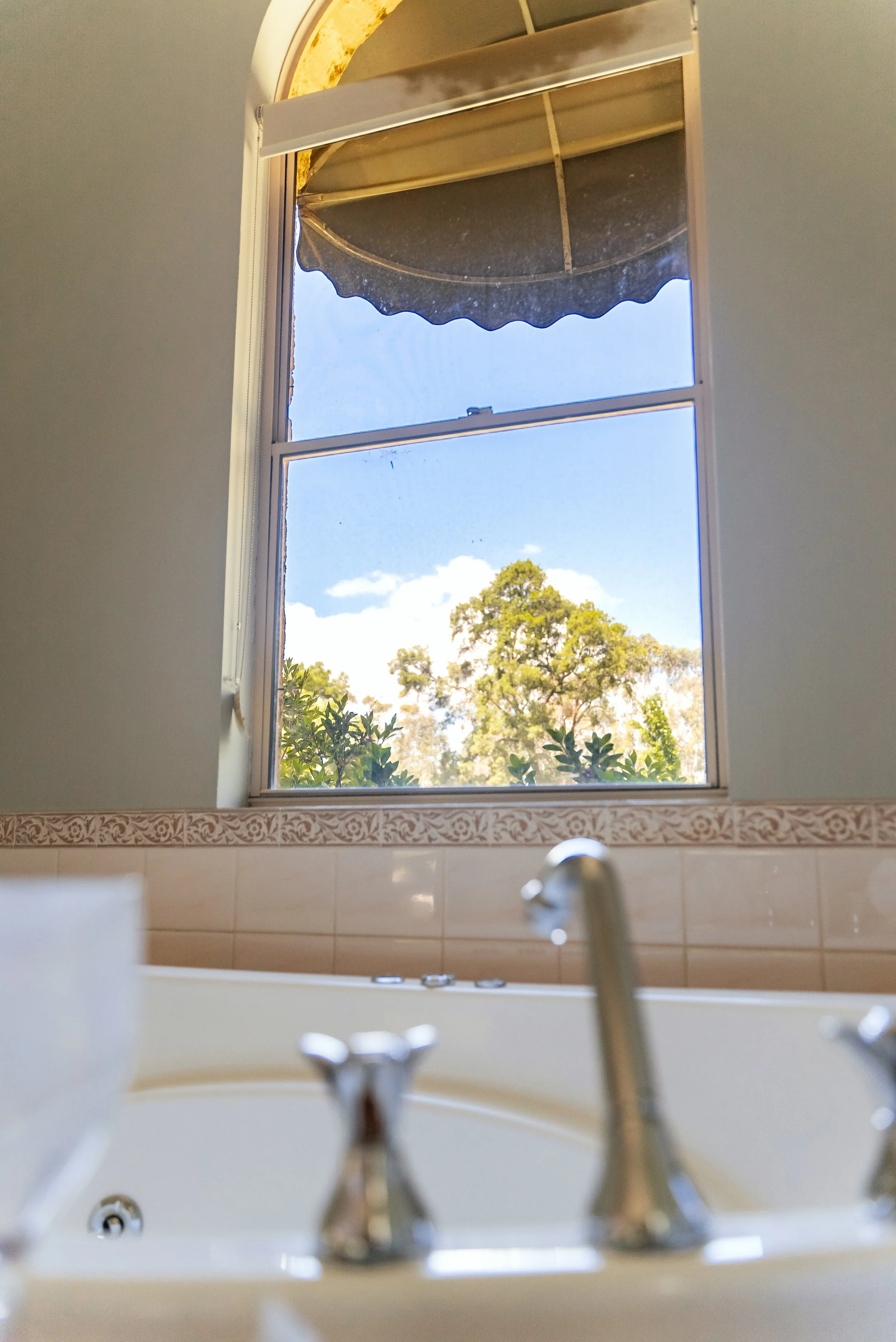 View of a bathroom with a bathtub in the foreground, a window above it, and trees outside with a blue sky and some clouds.