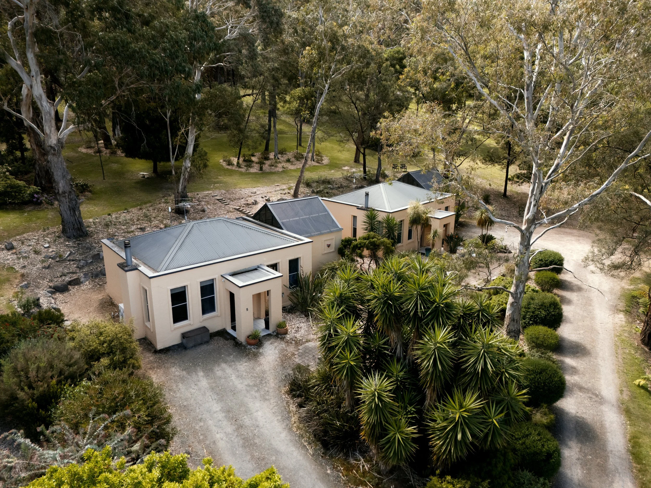 Aerial view of a modern single-story house surrounded by trees and bushes, with a gravel driveway and a lush green landscape nearby.