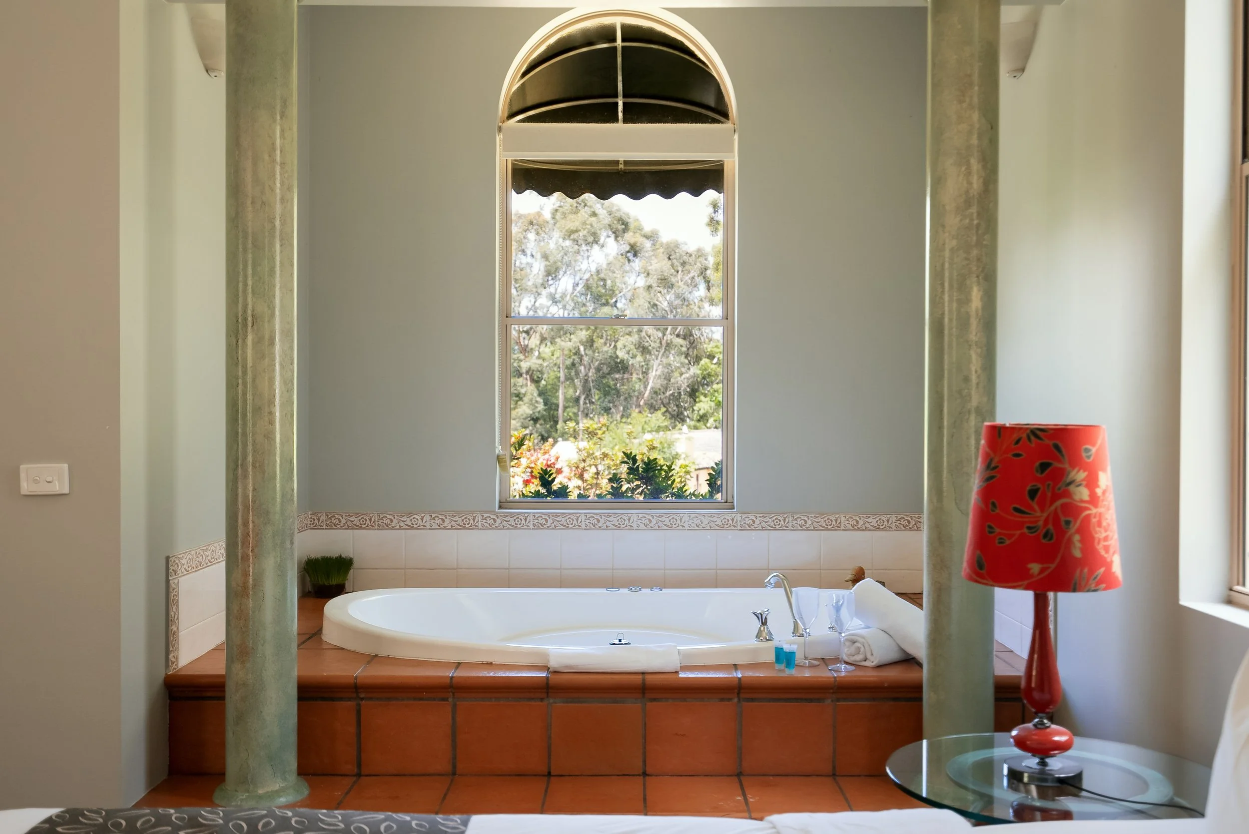 A bathroom with a large bathtub under a window, featuring a red lamp on a glass side table.