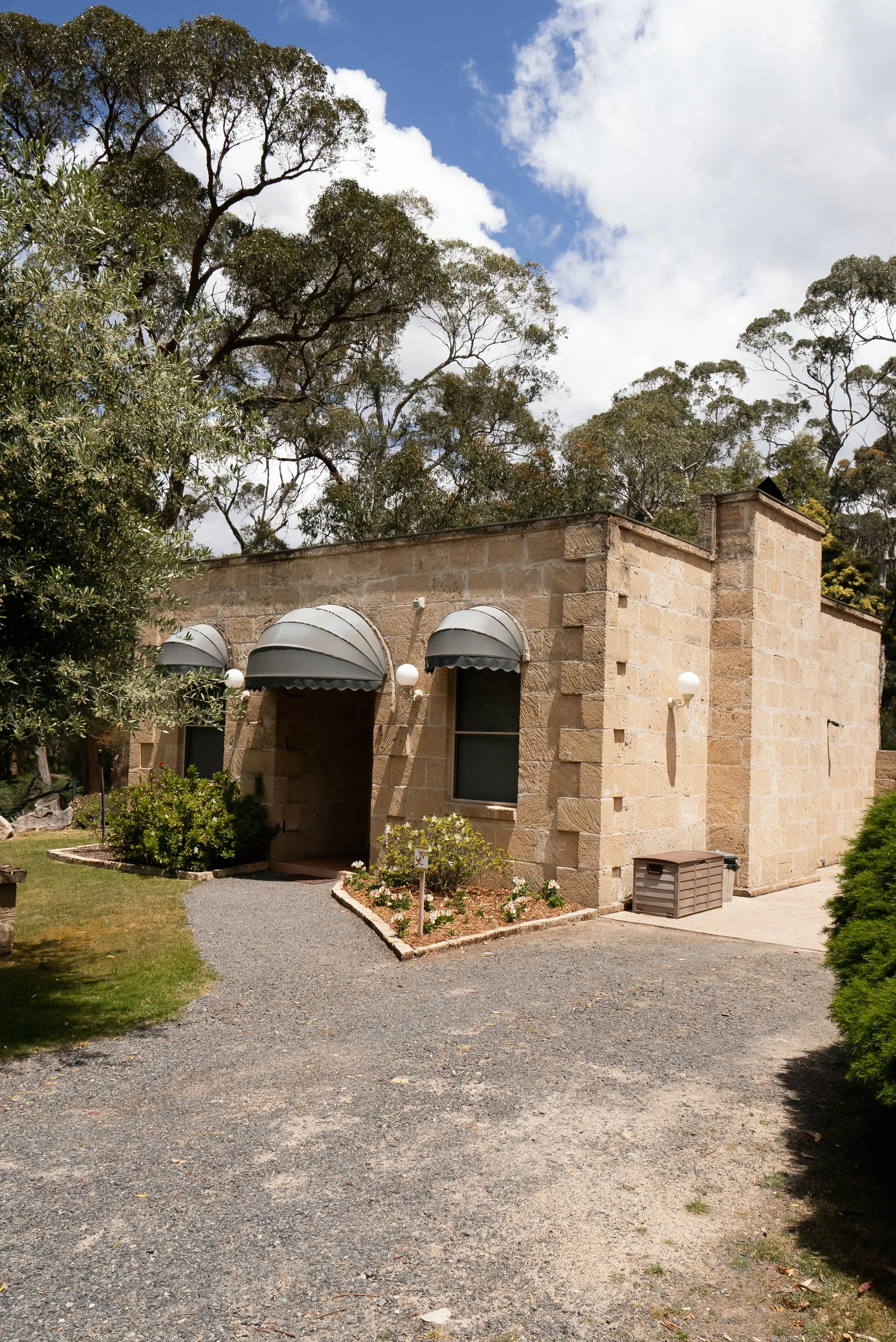 A stone building with three arched windows, each with a metal awning, surrounded by trees and a small garden with flowers and shrubs, under a partly cloudy sky.
