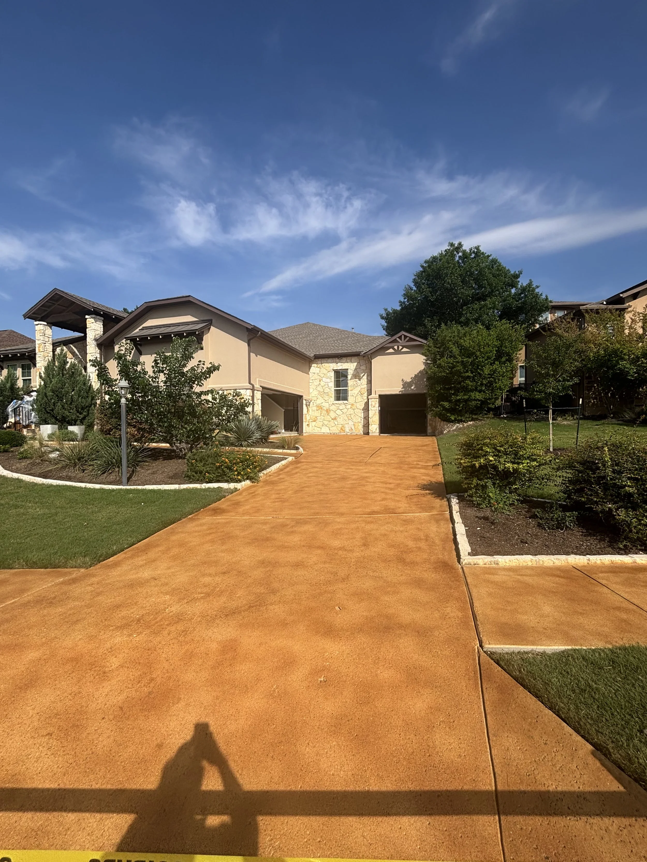A modern house with a brown concrete driveway leading up to two garage doors, surrounded by landscaped greenery and trees under a blue sky with some clouds.