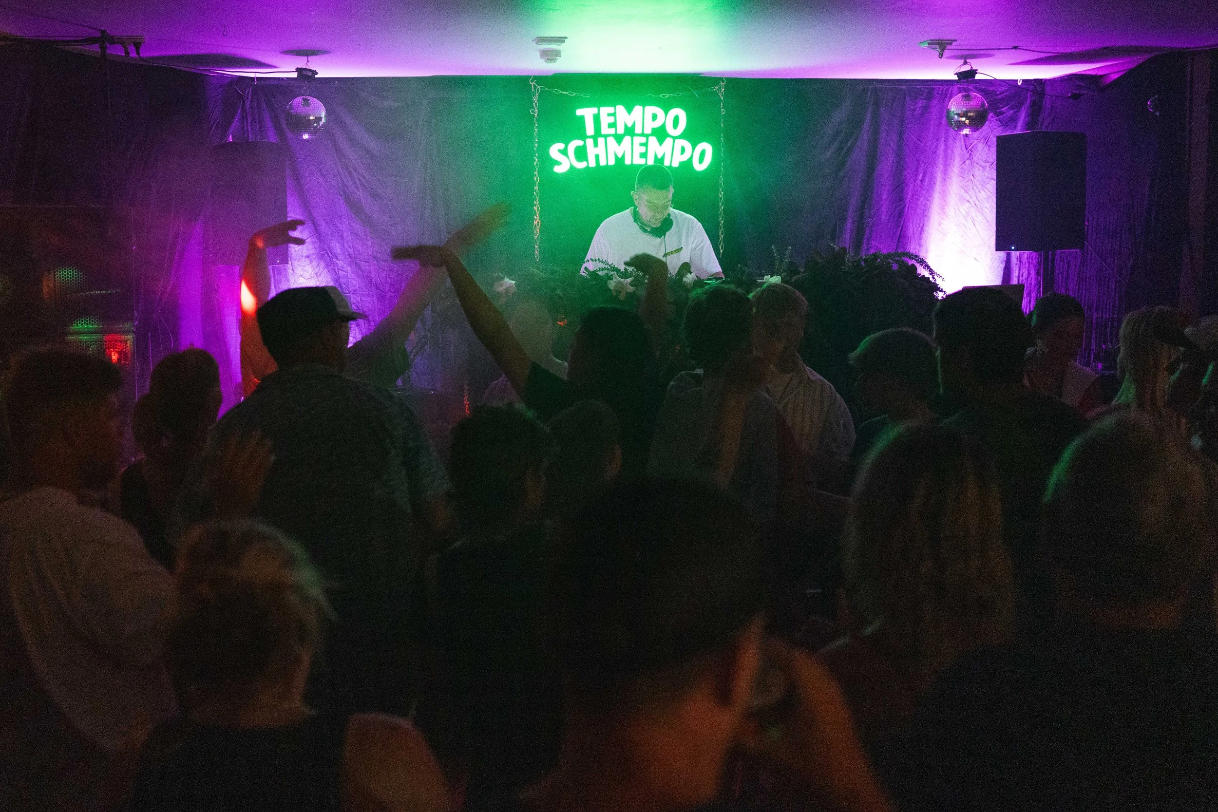 A crowd watching a DJ perform on stage with a neon sign that reads 'TEMPO SCHMEMPO,' colored purple and green lights, and people dancing.
