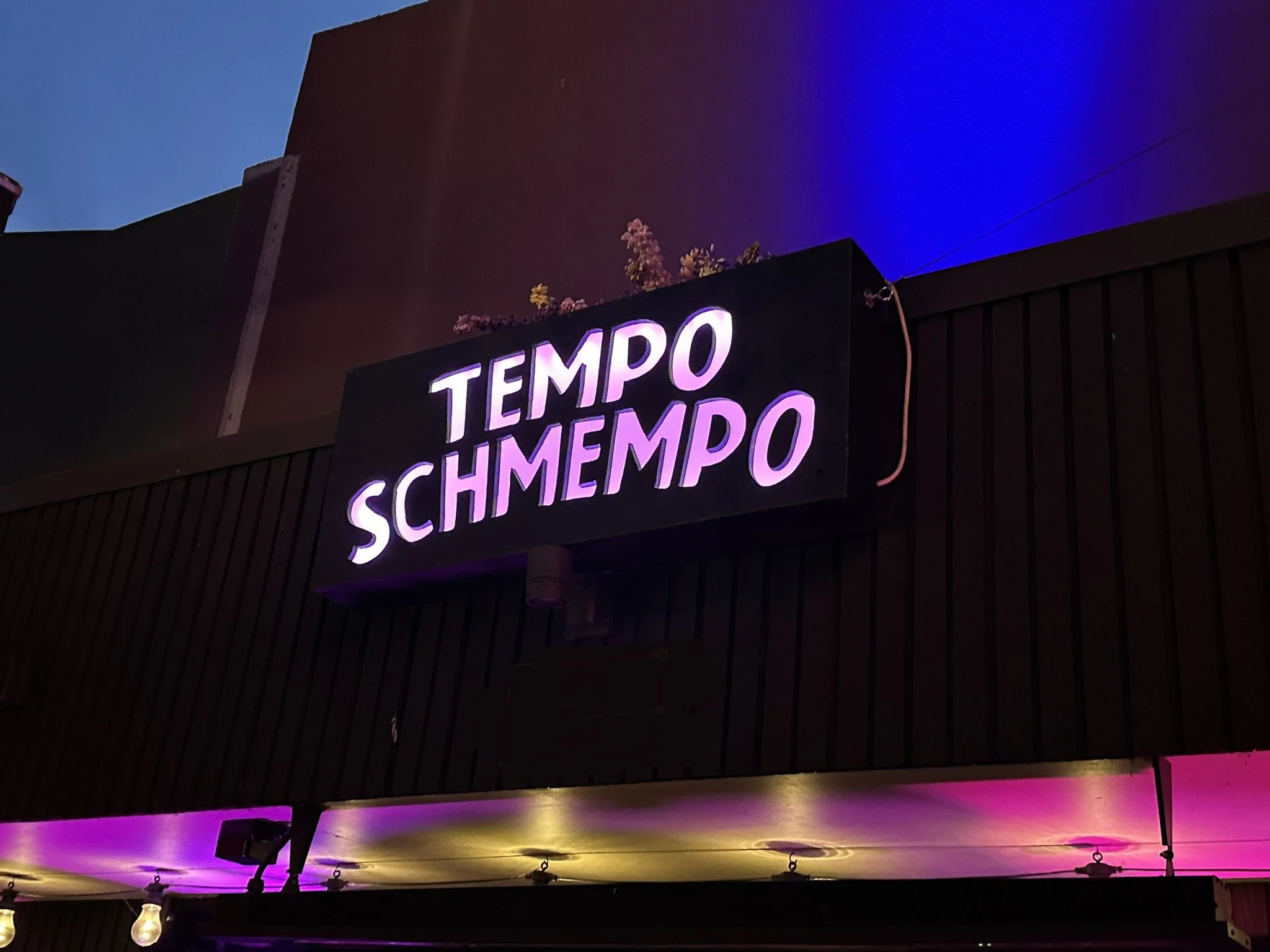 Neon sign reading 'TEMPO SCHMEMPO' in purple and white lights outside a building at night.