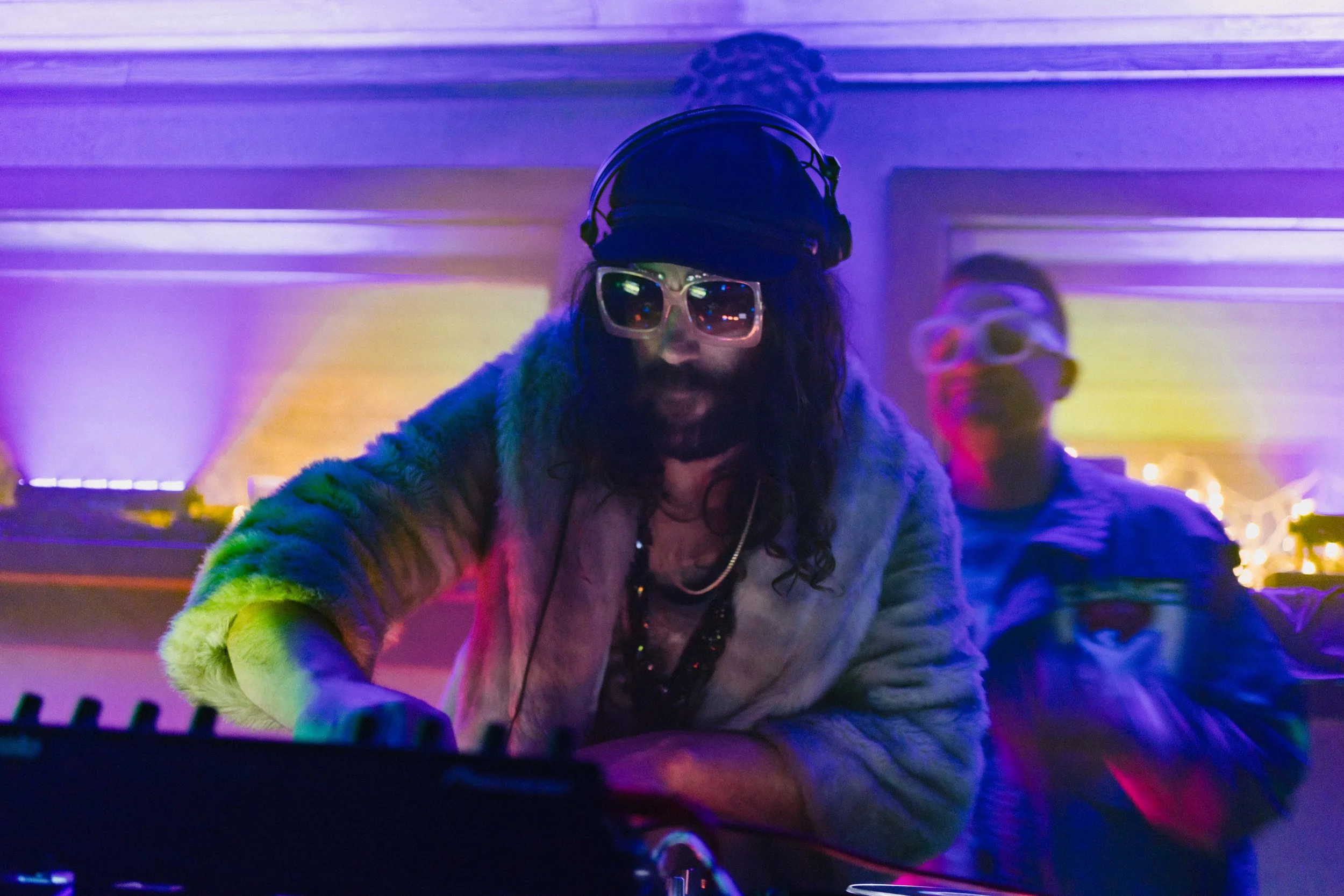 A DJ wearing large sunglasses, a black cap, and a fur jacket, performing at a night event with colorful lighting and decorations, with a man in sunglasses in the background.