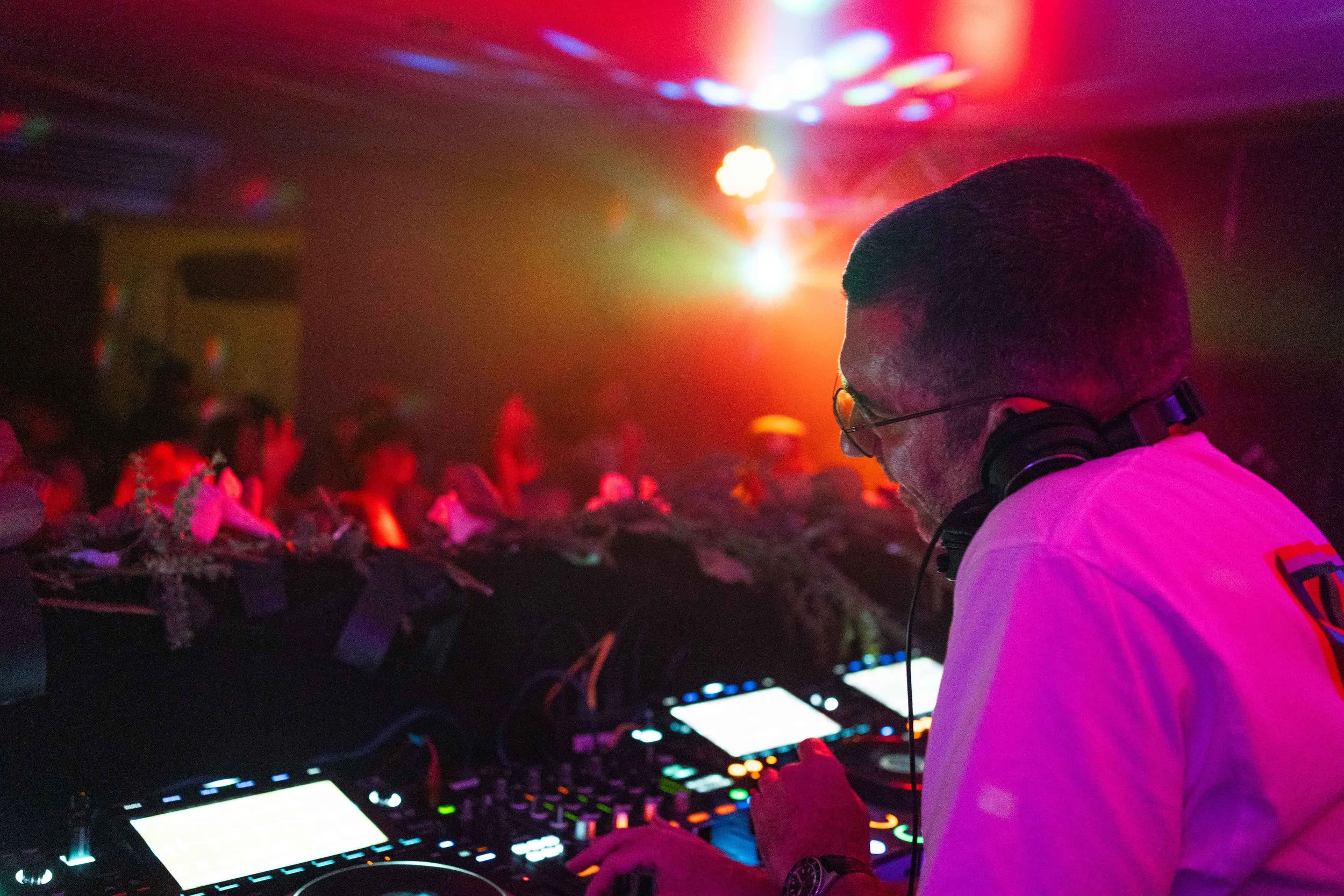 A DJ wearing glasses and headphones is performing at a nightclub with colorful lighting, and a crowd dancing in the background.