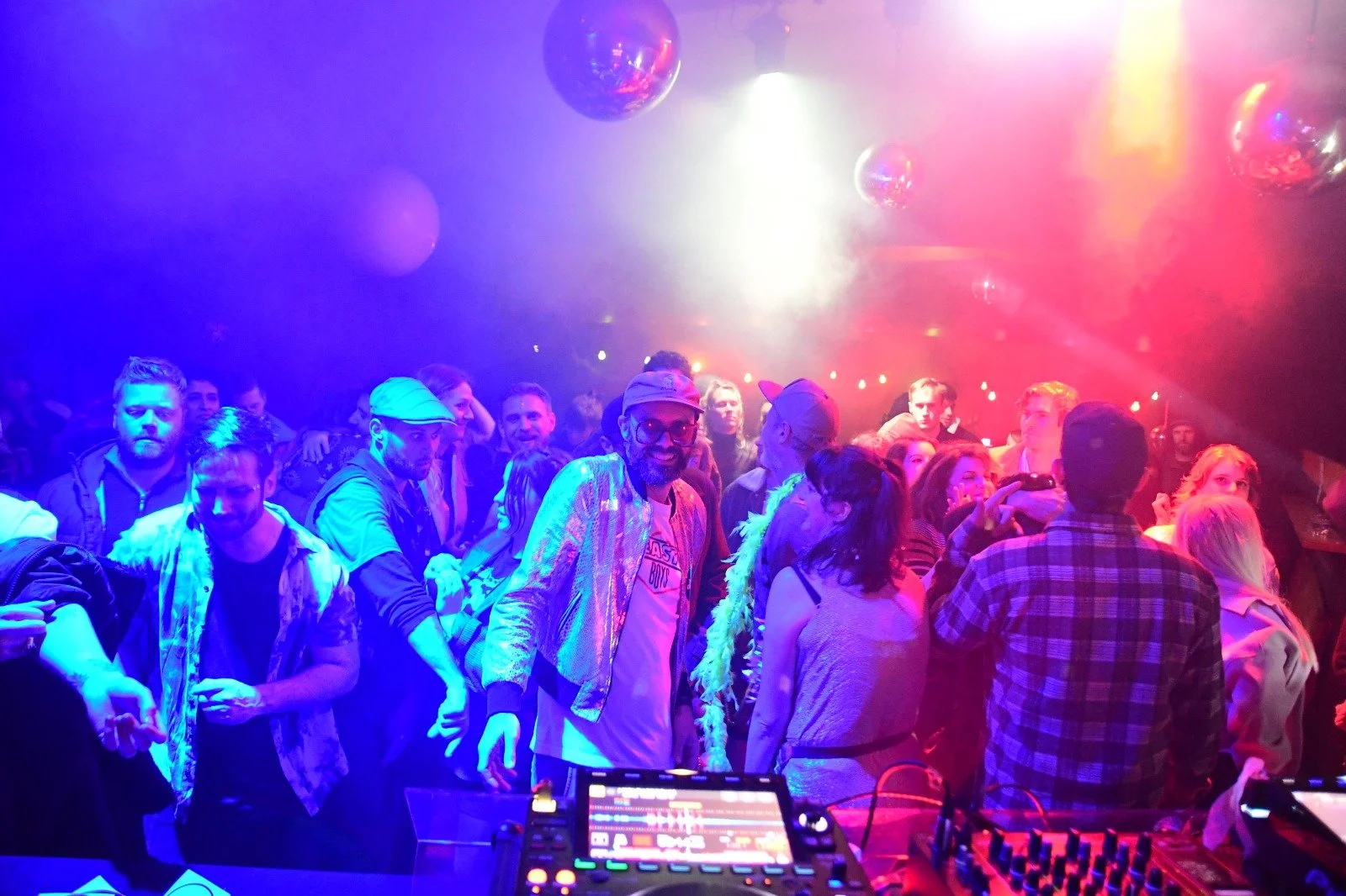 People dancing and socializing in a nightclub with colorful lights and mirrored disco balls overhead.