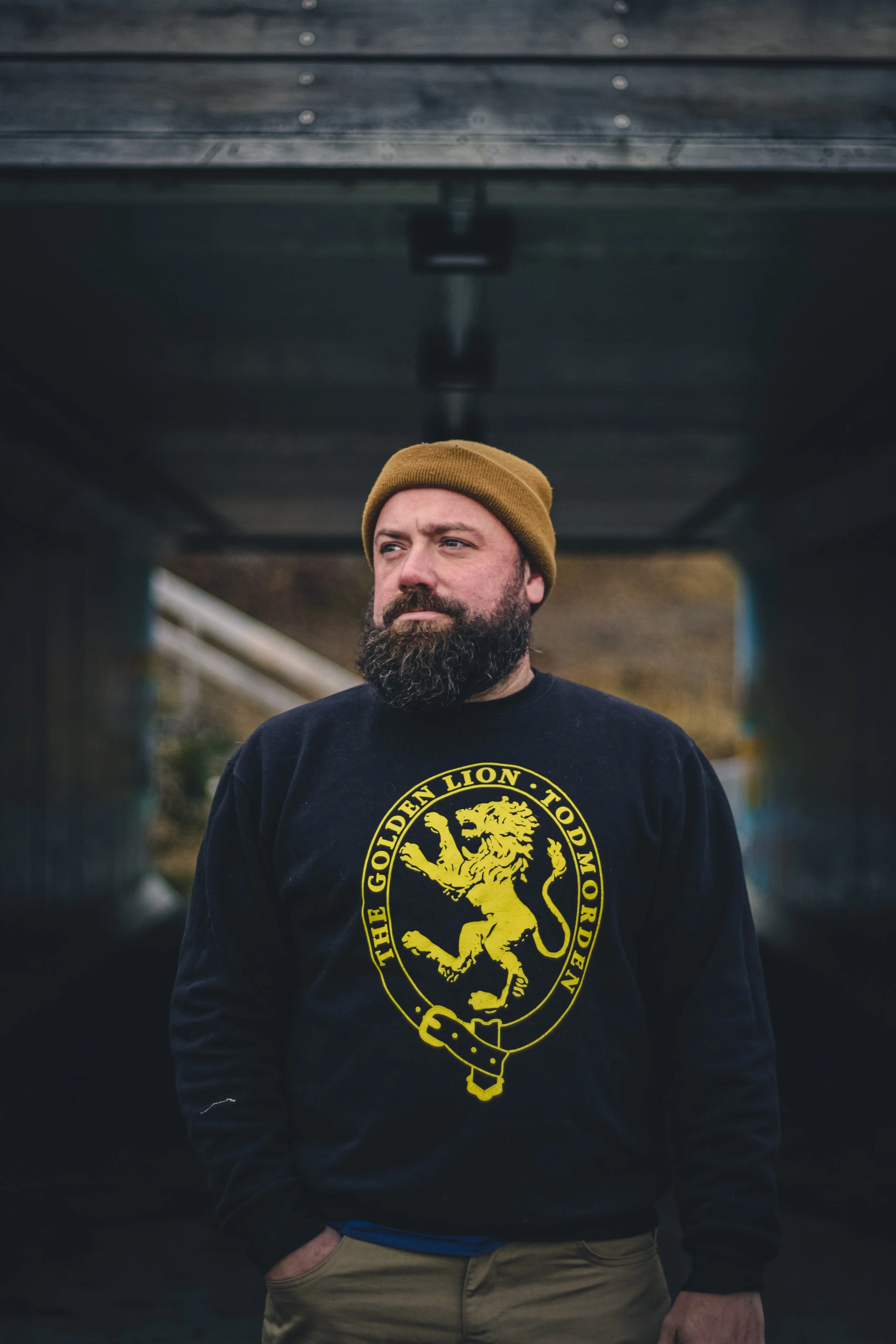 A man with a beard wearing a yellow beanie and a black sweatshirt with a yellow lion emblem in the center, standing outdoors. The background is blurred, and he appears to be under a bridge or similar structure.