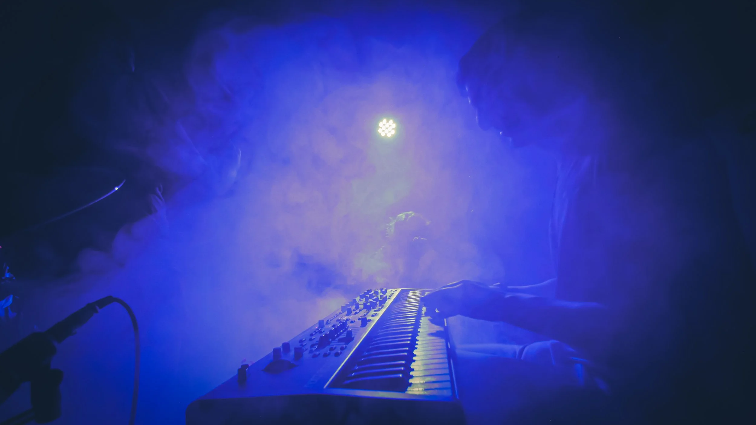 Two people playing a keyboard on a dimly lit stage with blue lighting and fog.
