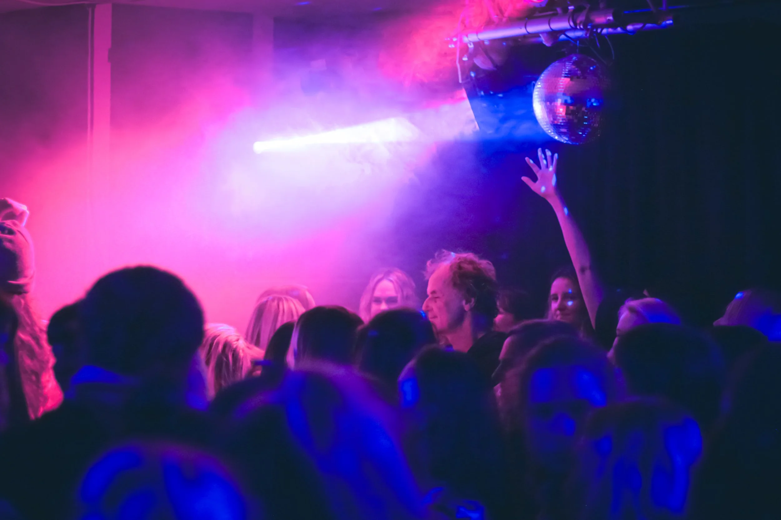 Crowd of people dancing in a nightclub with pink and blue lights, and a disco ball hanging from the ceiling.