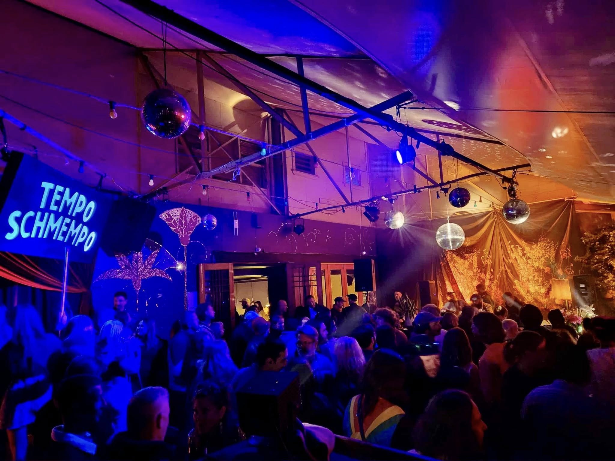 A lively indoor dance party with multicolored lighting, disco balls, and a crowd of people dancing and socializing.