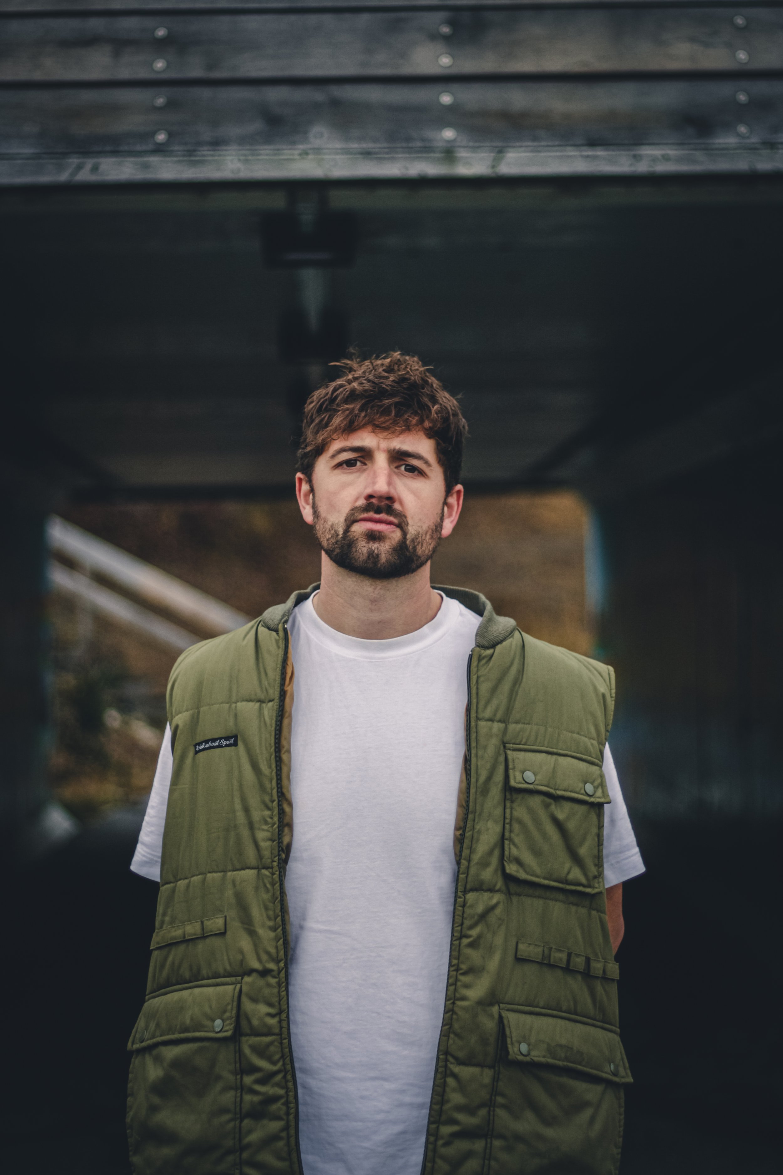 A man with a beard and brown hair standing under a bridge with a serious expression, wearing a white t-shirt and green vest.