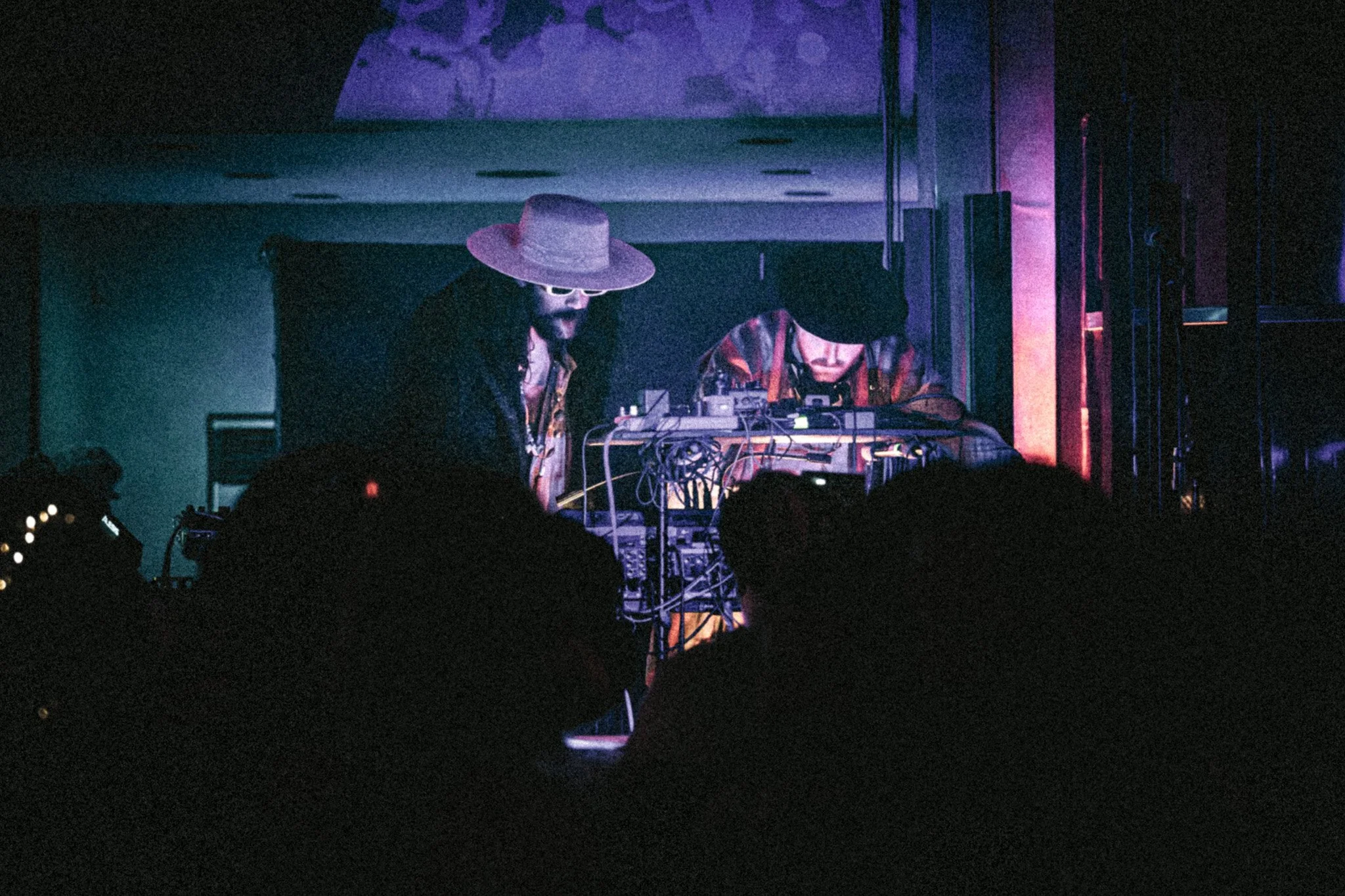 A DJ wearing a large straw hat and sunglasses performing on stage with a female DJ in a hat and glasses, surrounded by electronic music equipment, in a dimly lit venue.