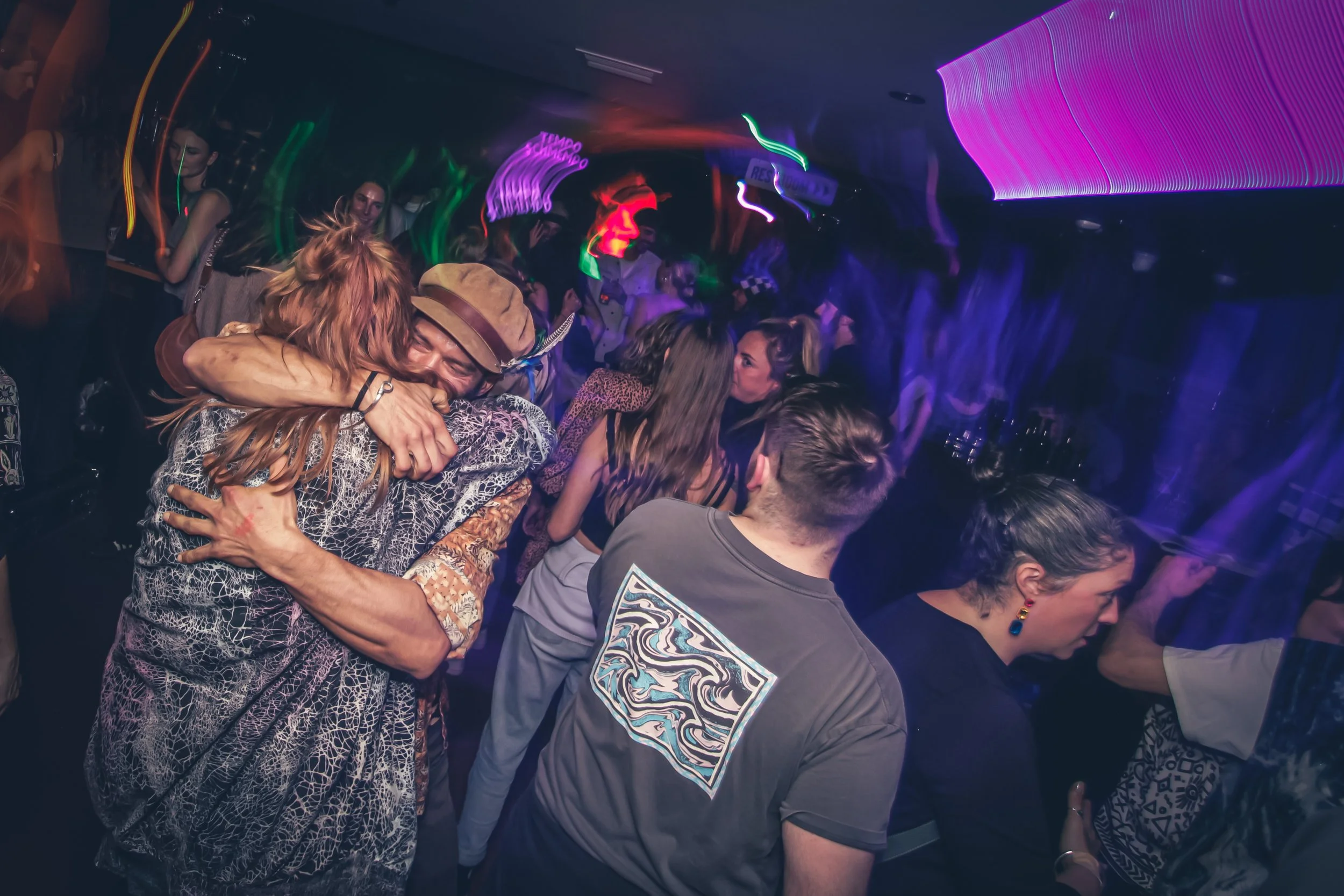 People at a nightclub dance floor, embracing and celebrating in colorful lights with motion blur.