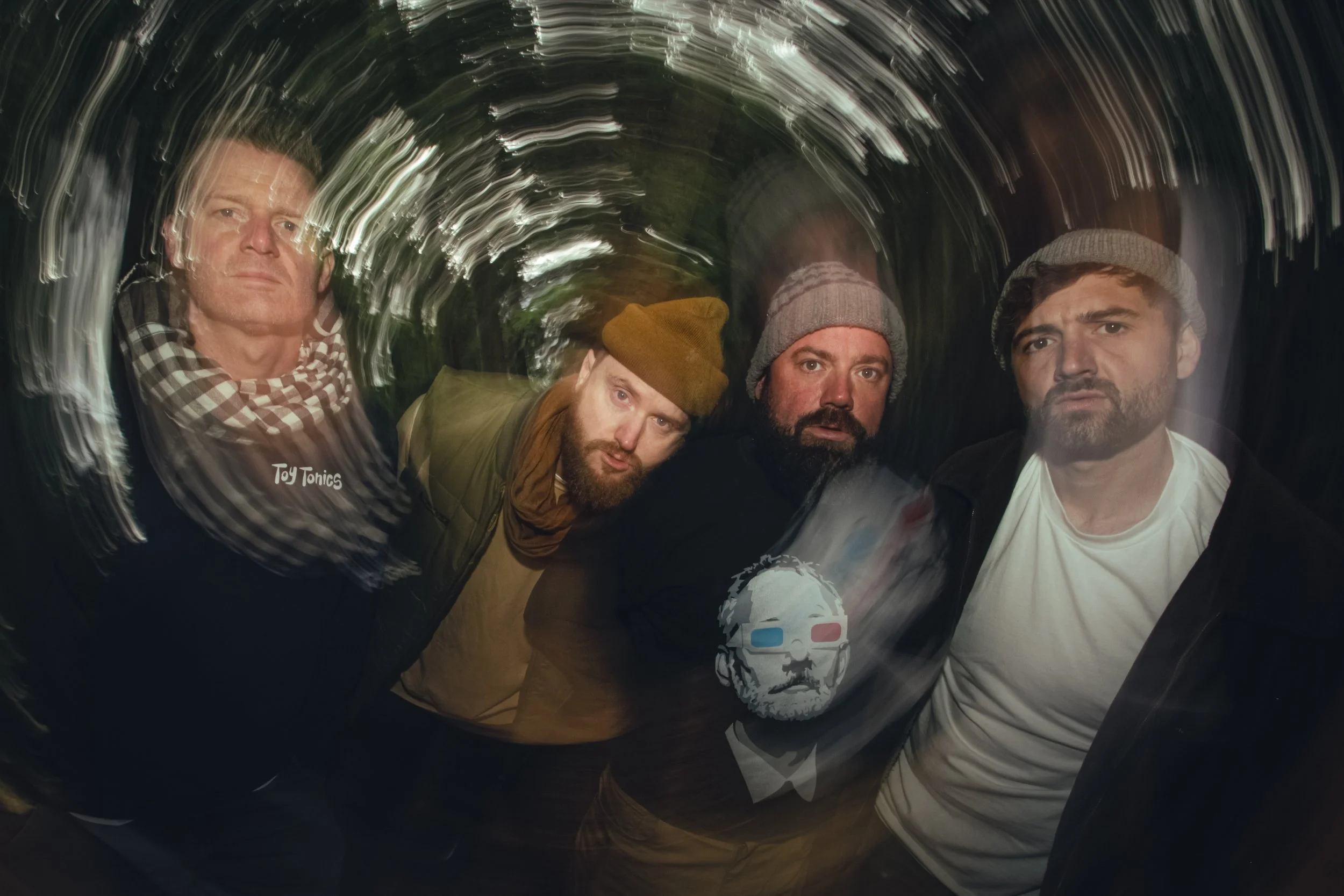 Four men with beards and hats, posing for a photo with a swirl effect background.
