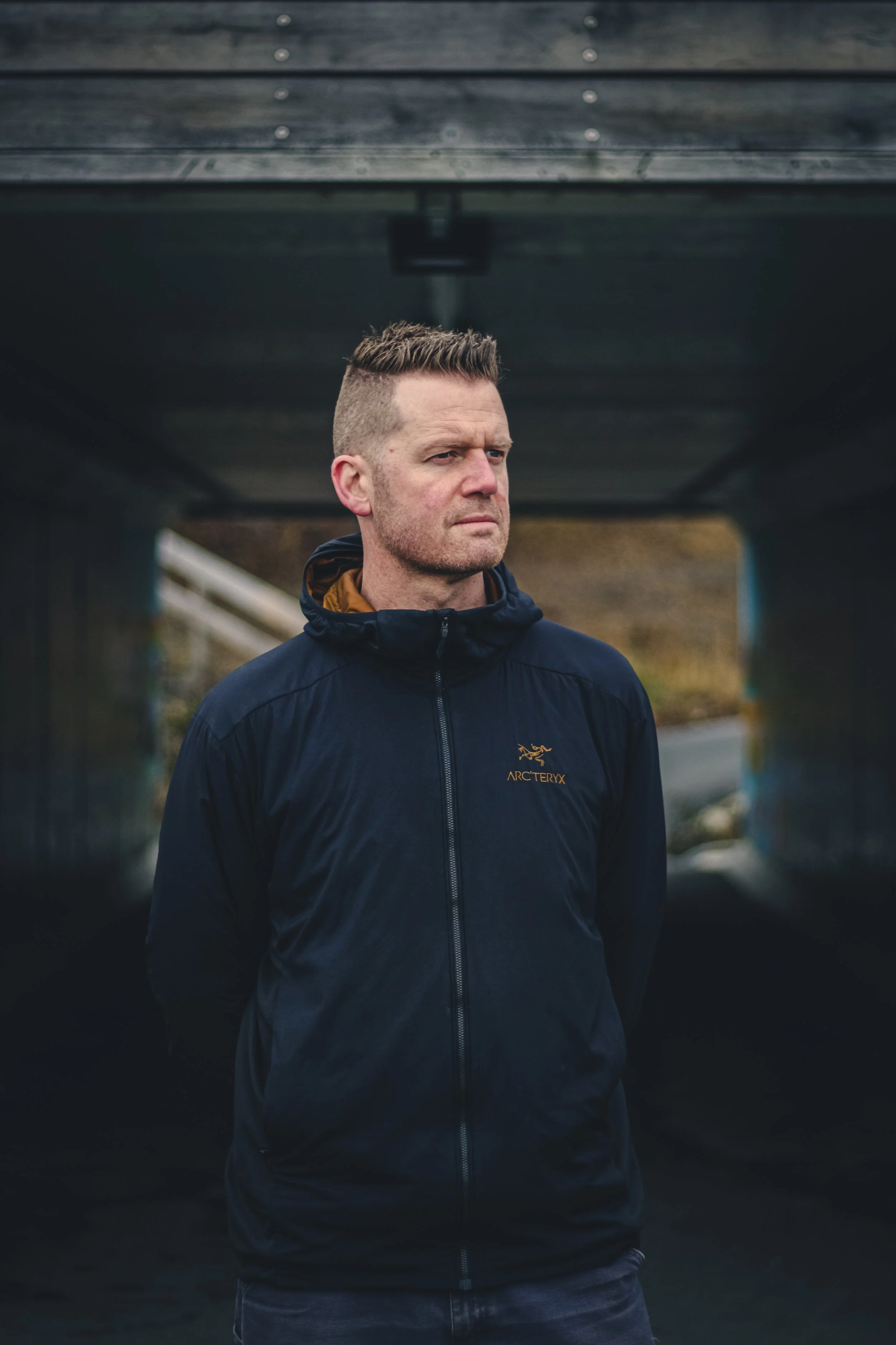 A man with short, styled hair wearing a navy Arc'teryx jacket standing under a bridge on a cloudy day.