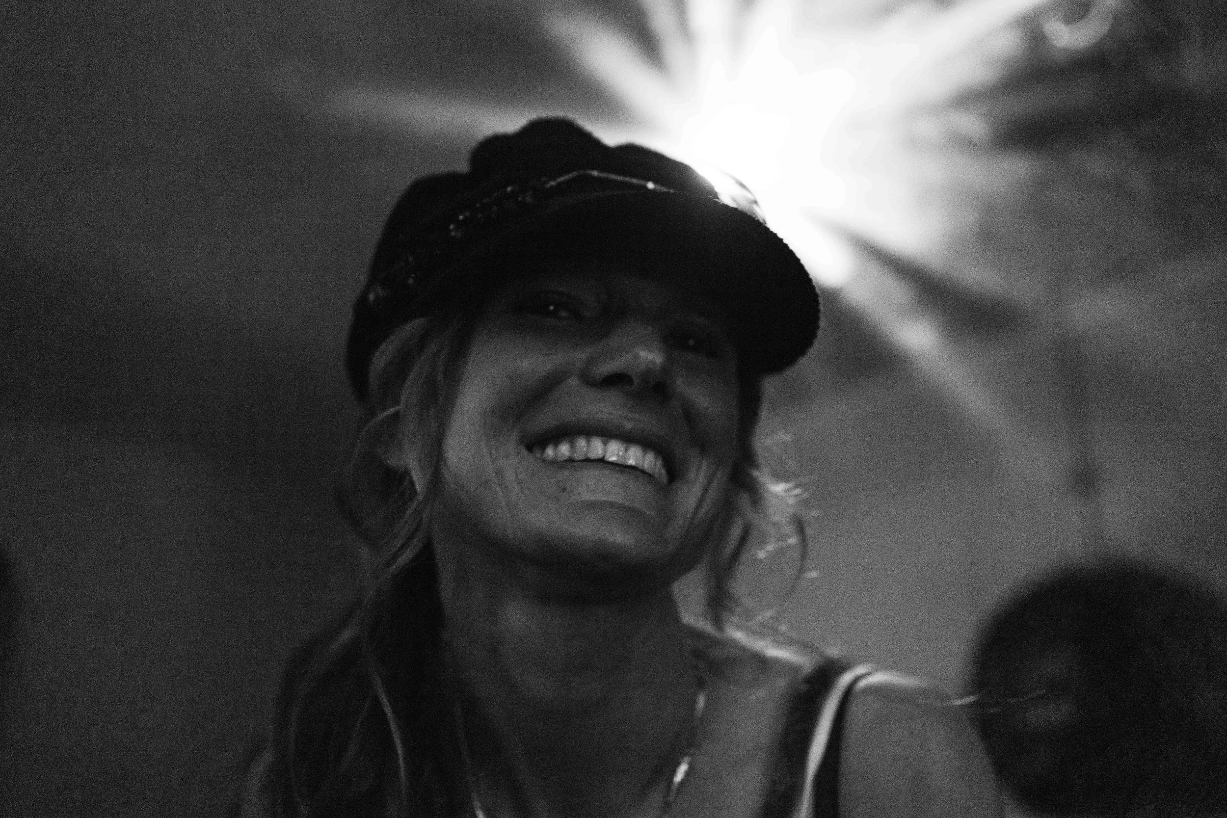 A woman smiling, wearing a cap and tank top, in a dark setting with a bright light behind her head.
