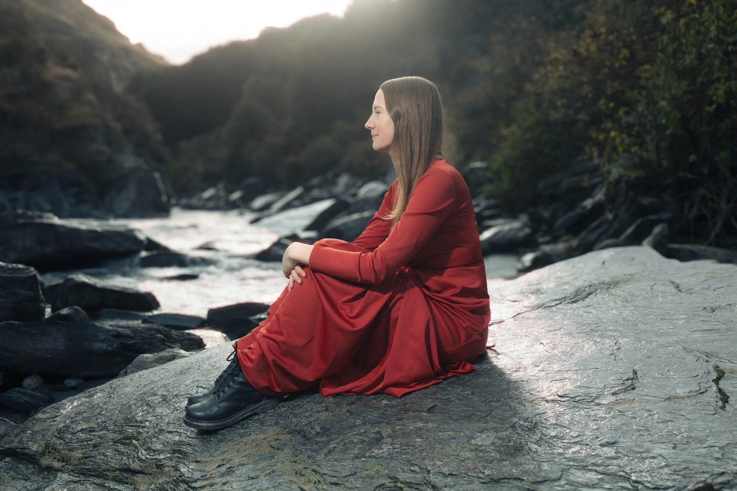 Woman sitting on a large rock by a river, dressed in a red dress and black boots, with a forested canyon in the background.