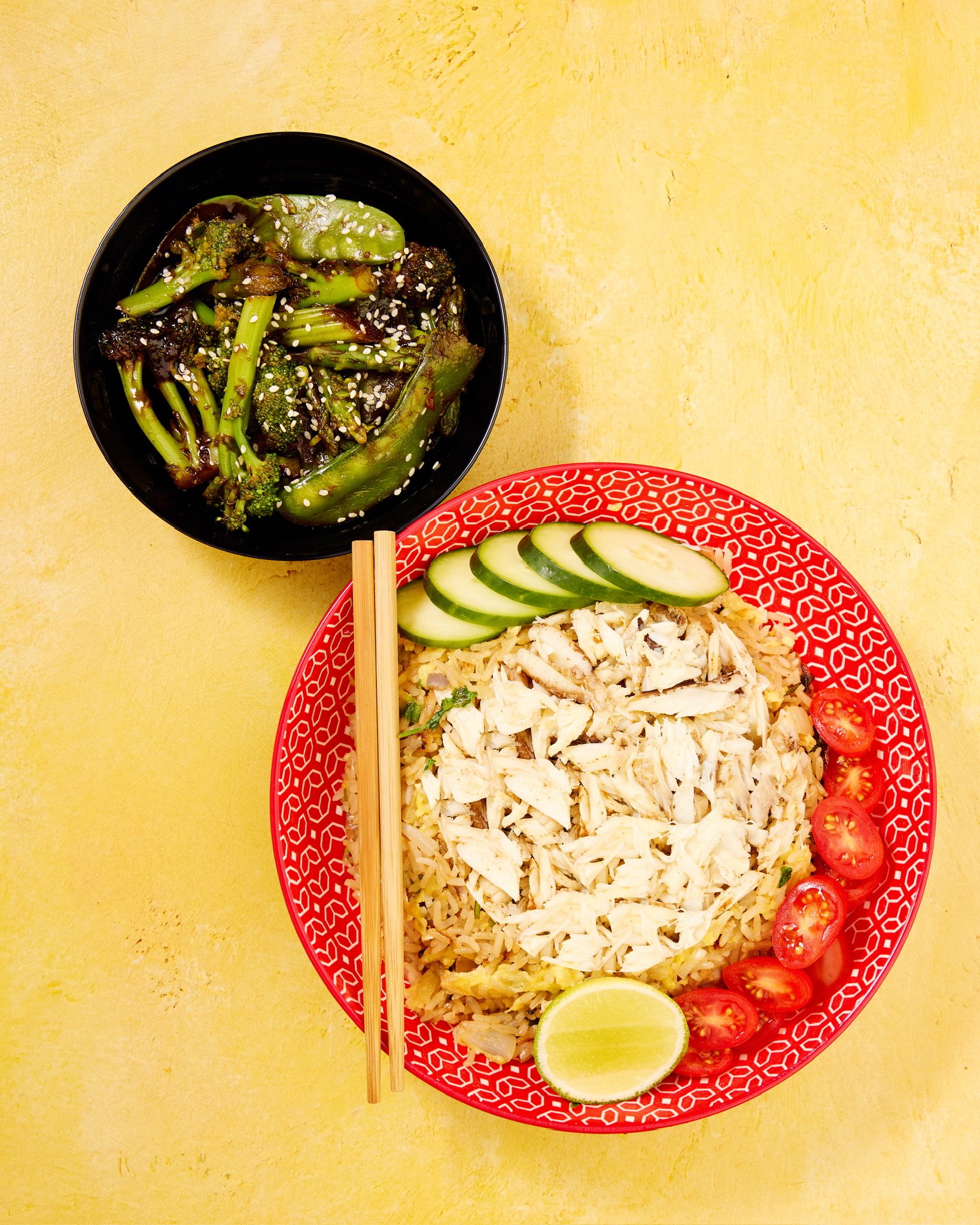 Asian-style meal with rice topped with crabmeat, cucumber slices, cherry tomatoes, lime wedge on a red patterned plate, and a bowl of sautéed broccoli and snap peas with sesame seeds on a yellow background.