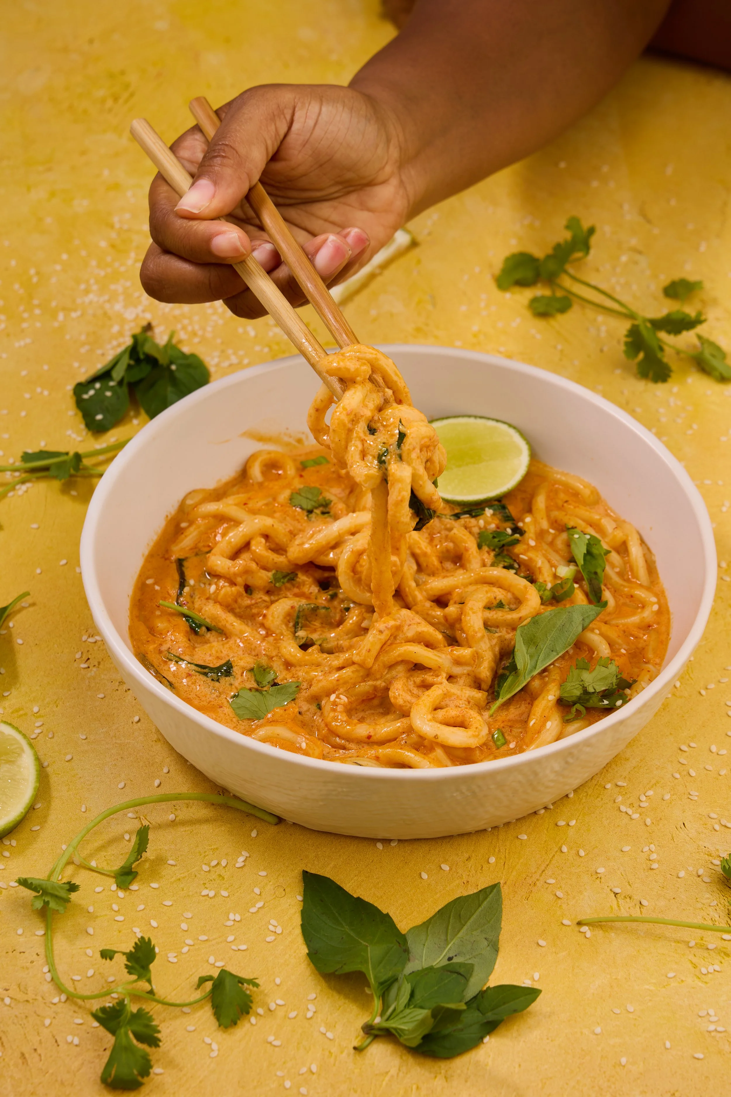 Person using chopsticks to pick up noodles from a bowl of curry noodles. The bowl sits on a yellow surface decorated with lime slices, cilantro, basil, and sesame seeds.