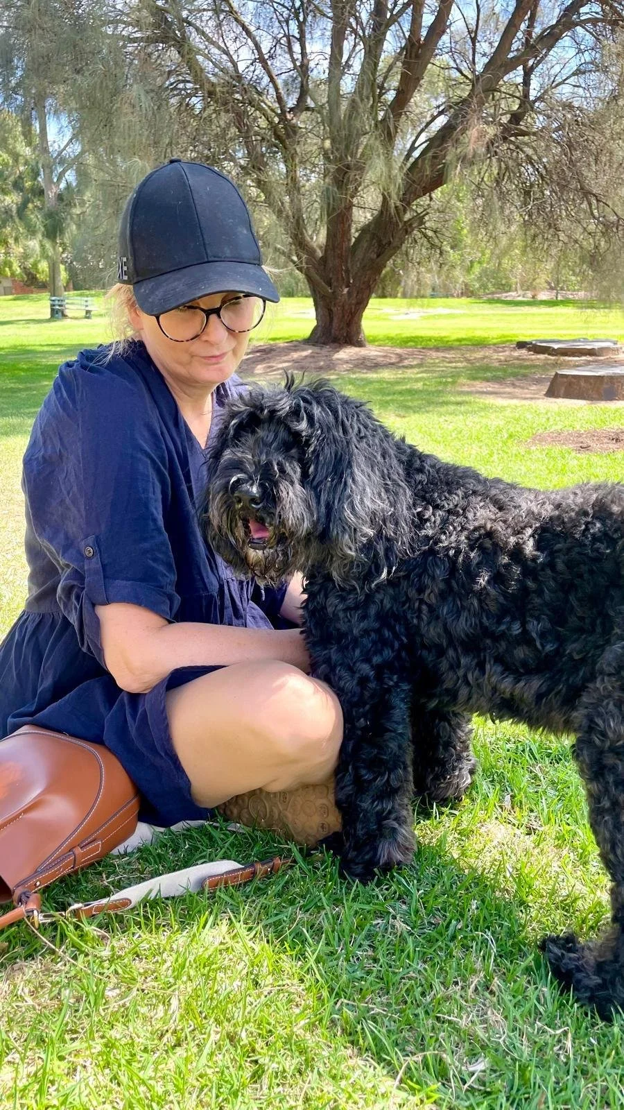 A woman wearing glasses, a black cap, and a navy blue dress sitting on grass in a park with a large black curly-haired dog. The woman is gently holding the dog, and they are both under a tree with a bright, sunny background.