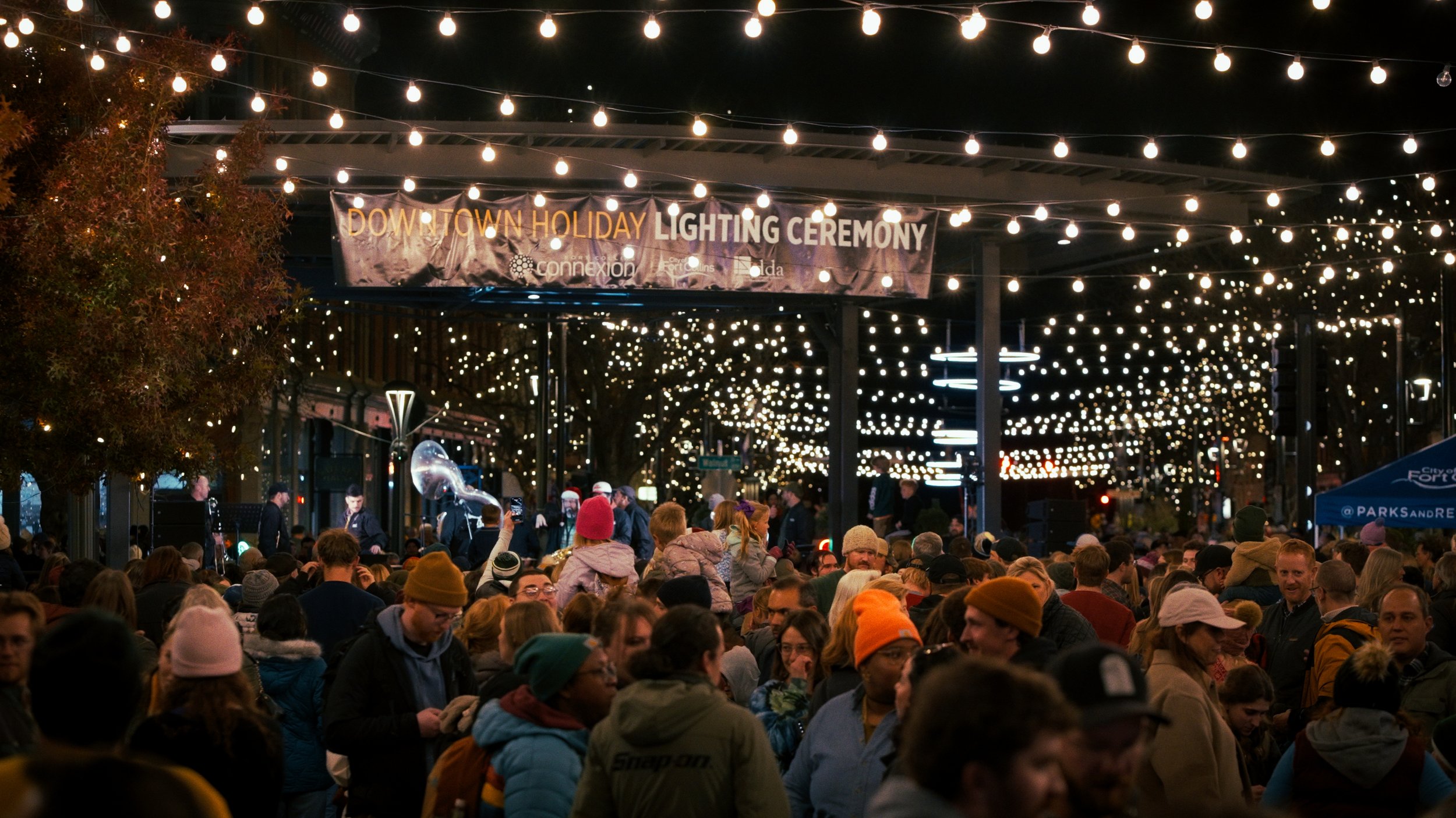 Crowd gathered at a nighttime downtown holiday lighting ceremony with string lights overhead and a banner that reads 'Downtown Holiday Lighting Ceremony'.