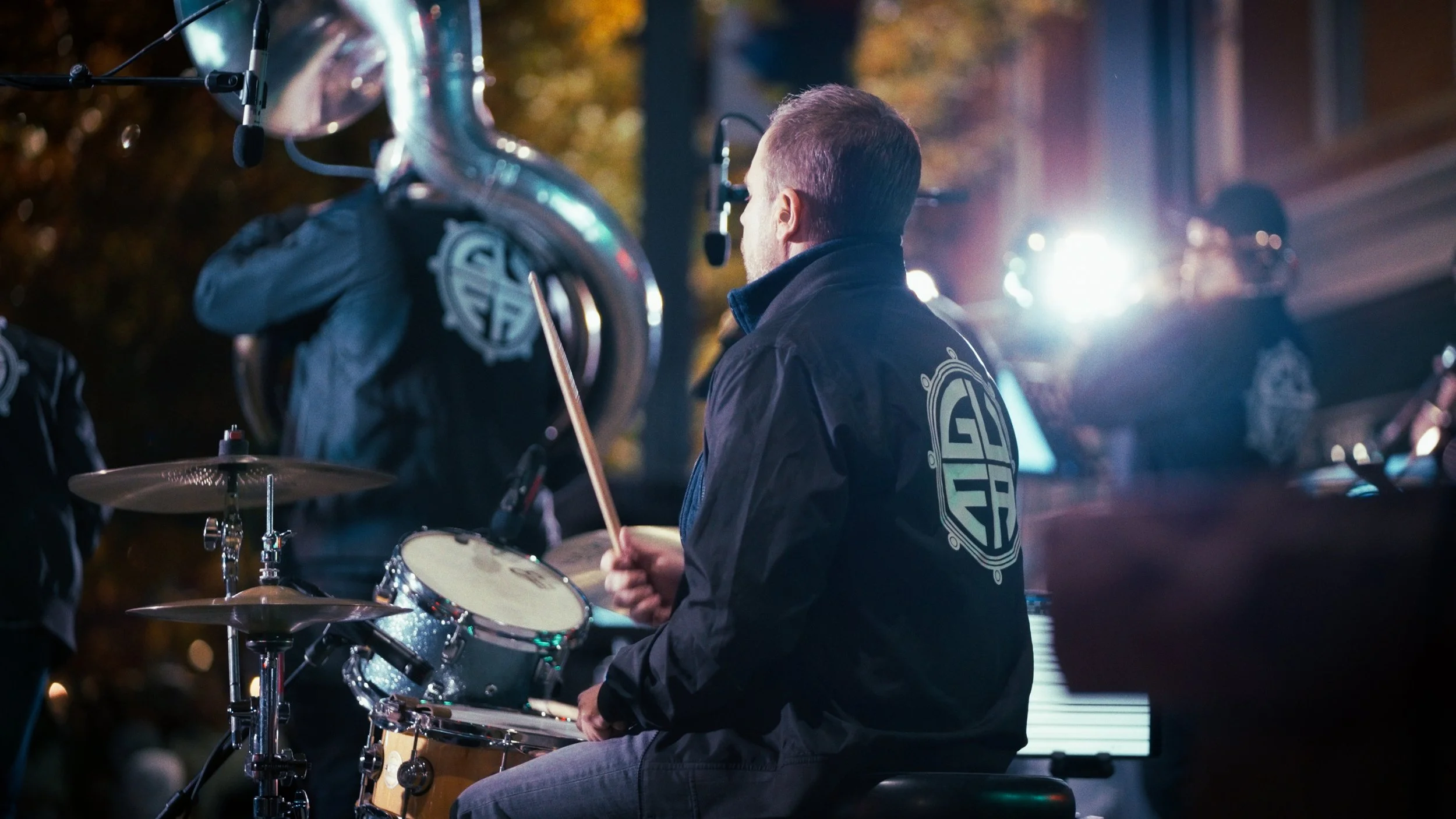 A man playing drums outdoors at night with bright lights in the background.