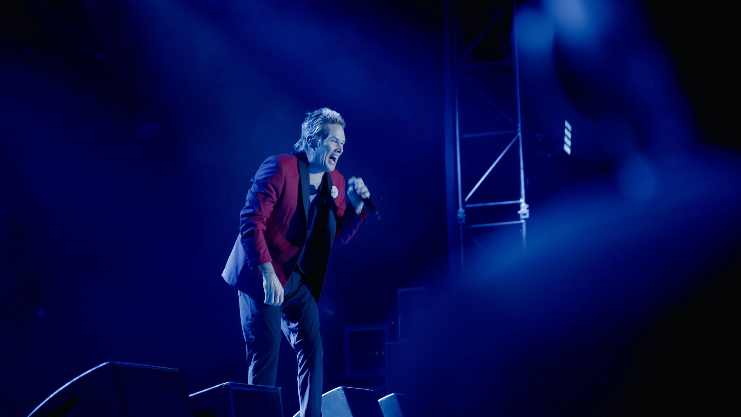 Male performer in a red blazer and gray pants on stage, smiling and holding a microphone, with blue stage lighting.
