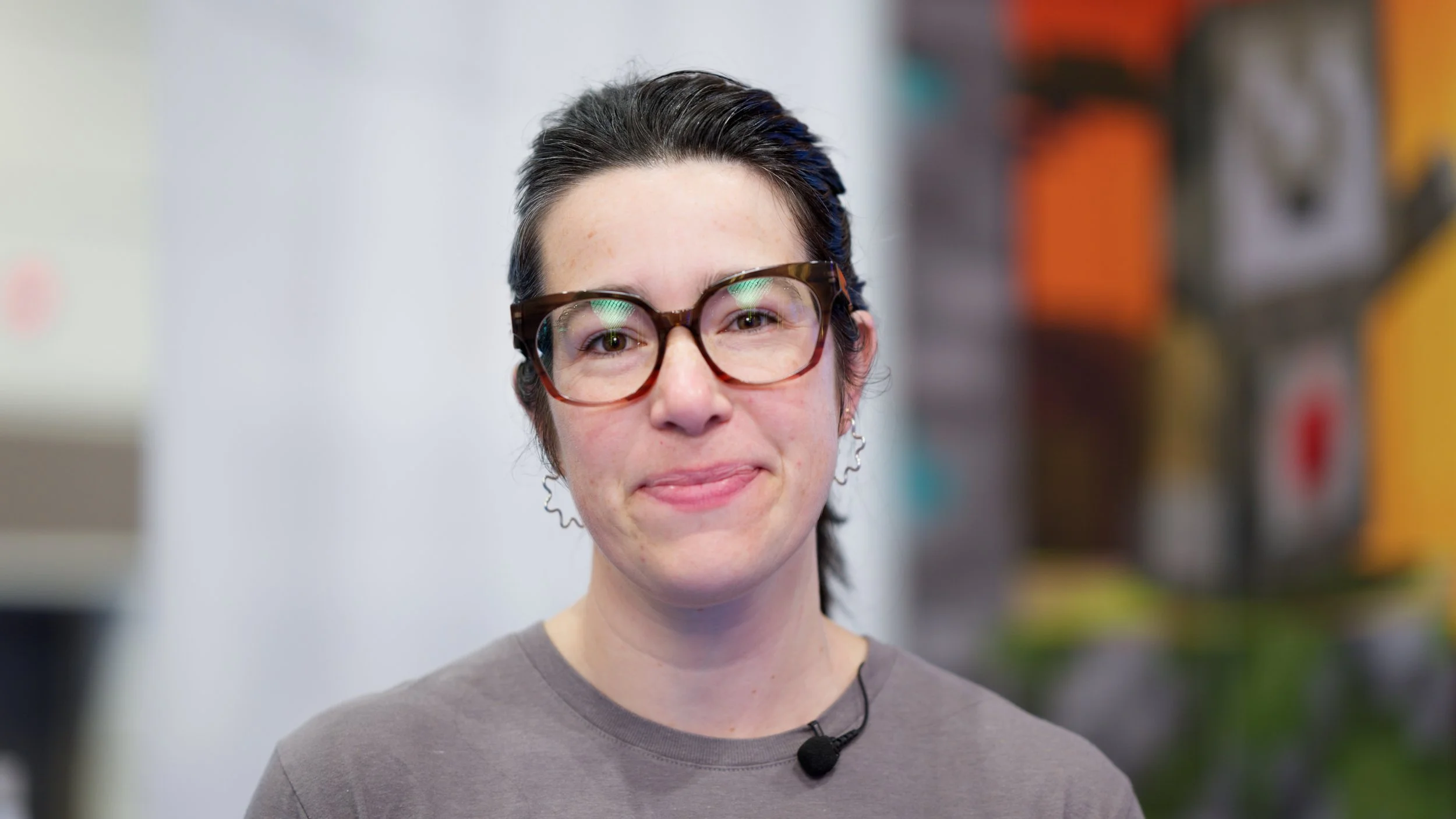 A young woman with short dark hair, wearing glasses and a gray shirt, smiling while looking at the camera.