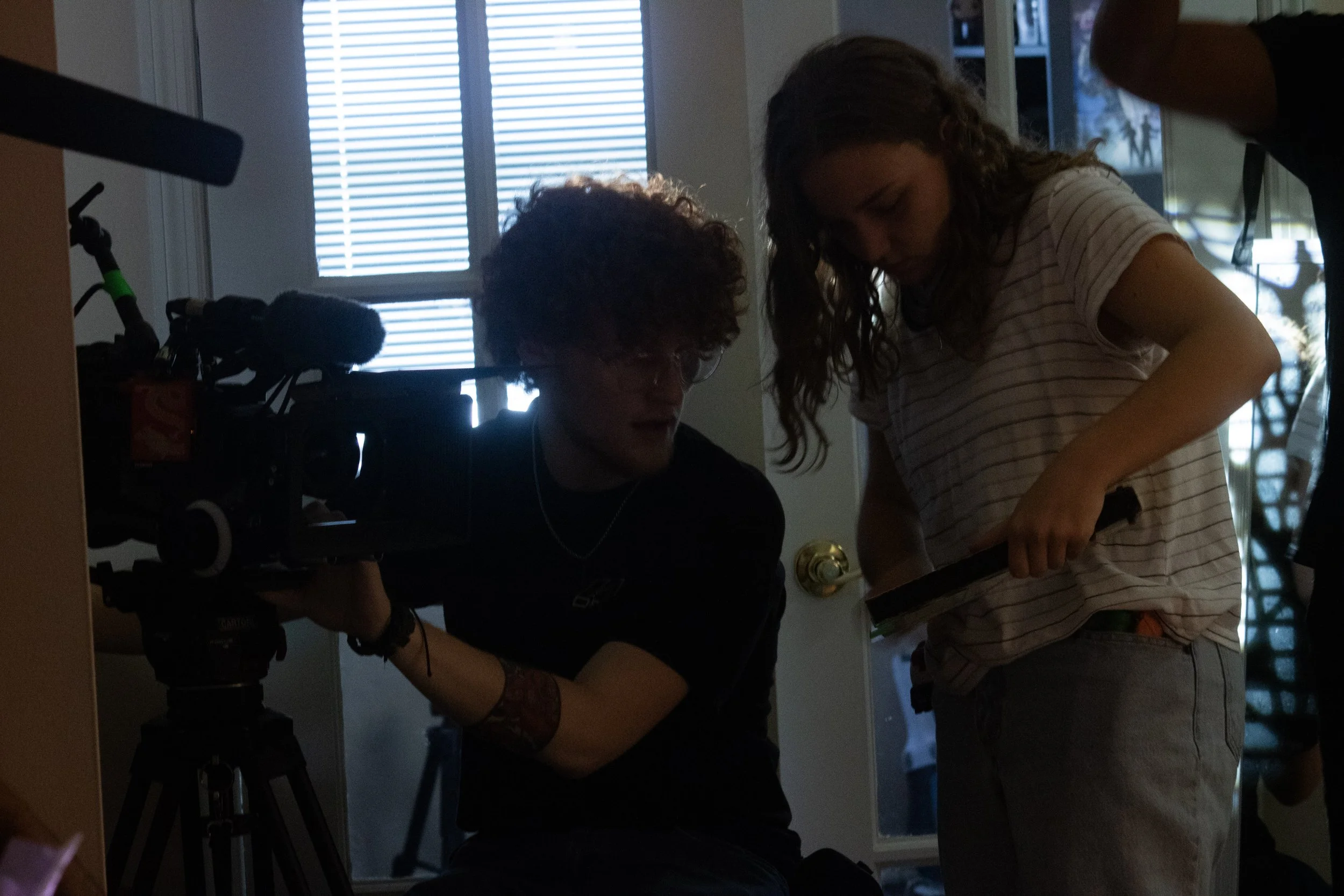 Two women working with film equipment in a room with a window with blinds. One woman is sitting and operating a camera, while the other woman is standing and adjusting something near her.