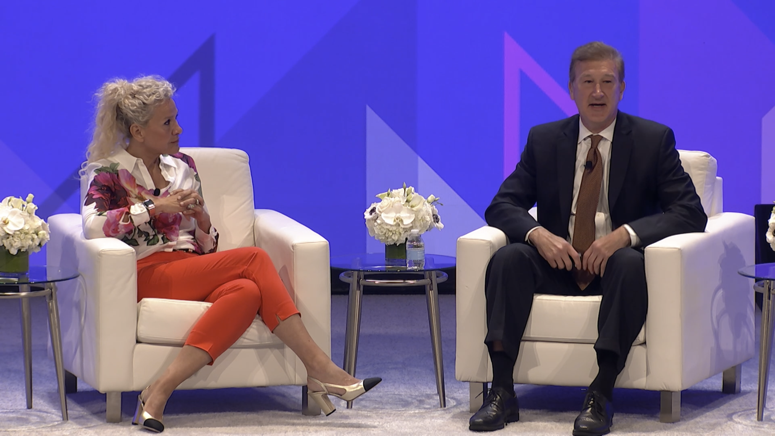 A woman and a man sitting on white armchairs on a stage, engaged in conversation. The woman has curly blonde hair, is wearing a white blouse with floral patterns, red pants, and beige heels. There are flowers in glass vases on small tables beside them, and a blue background with geometric patterns.
