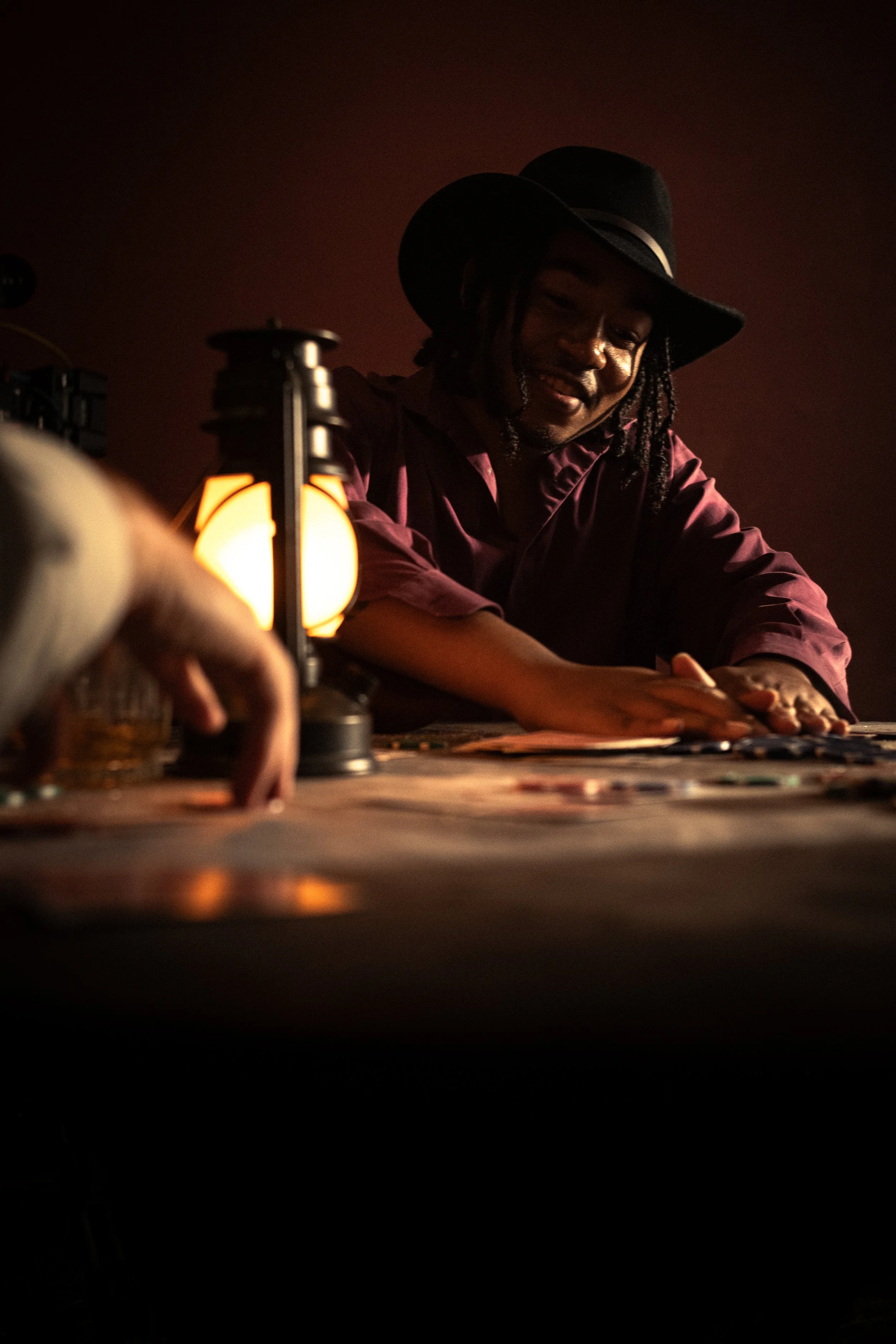 Man wearing a black hat with a brim, smiling, and seated at a table with hands resting on the surface. Warm lighting and a blurred lamp in the foreground.