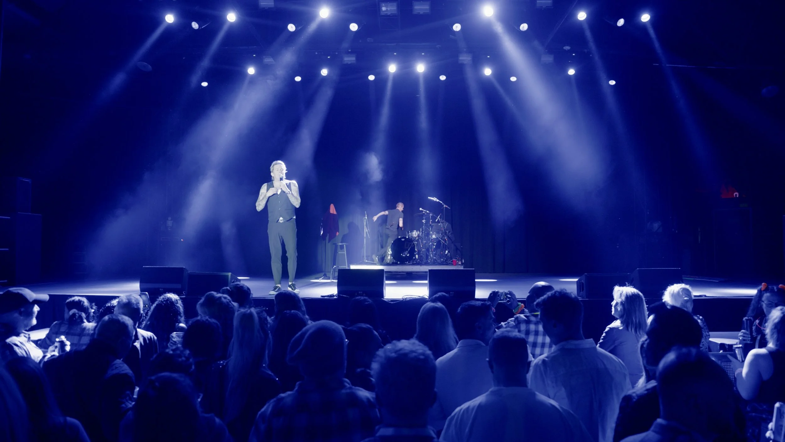 A singer performing on stage with a microphone, accompanied by band members and drums, in front of an audience at a concert venue with bright stage lights.