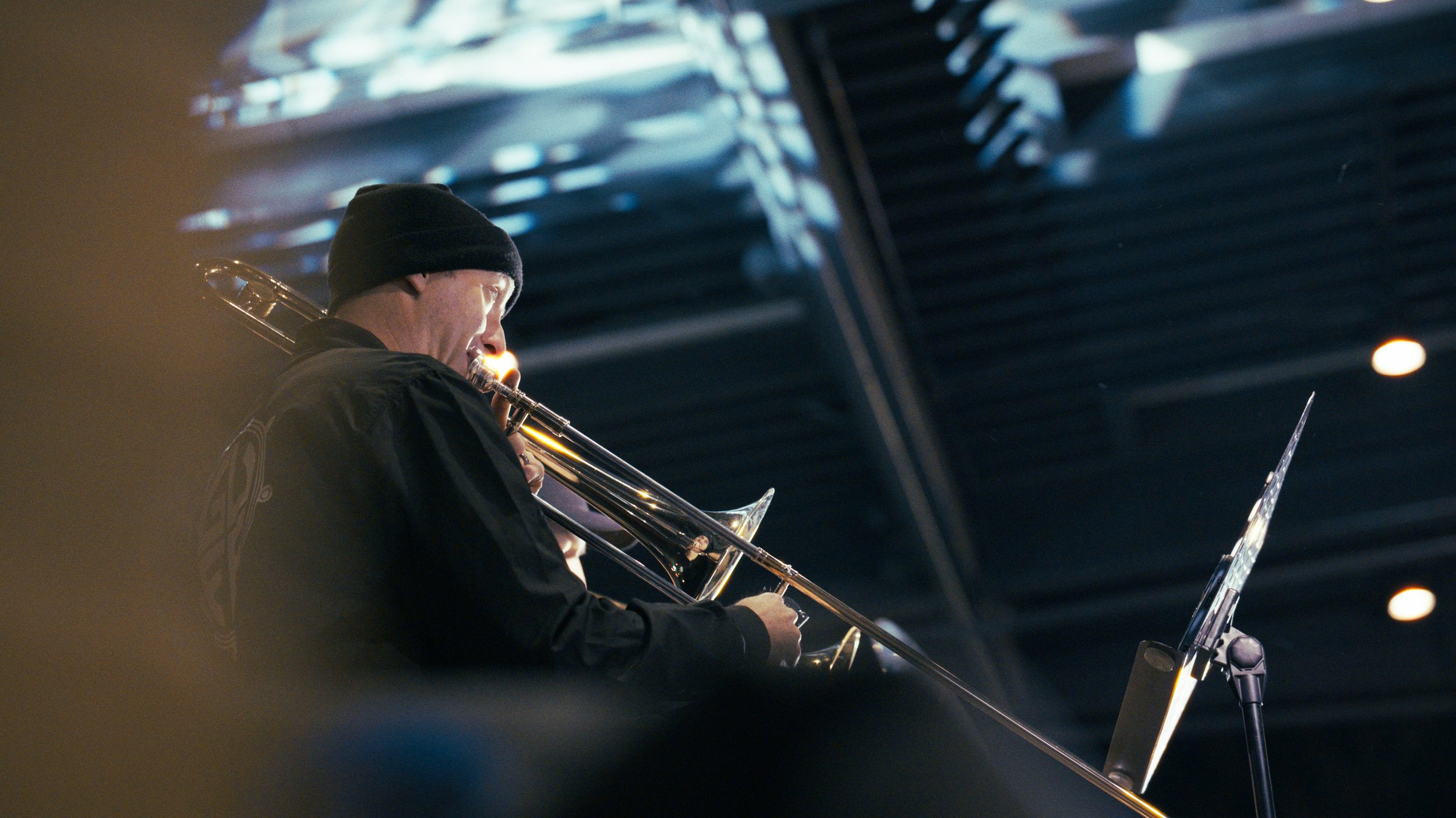 A man wearing a black beanie playing a trombone, with sheet music on a stand, under either stage or outdoor lighting.