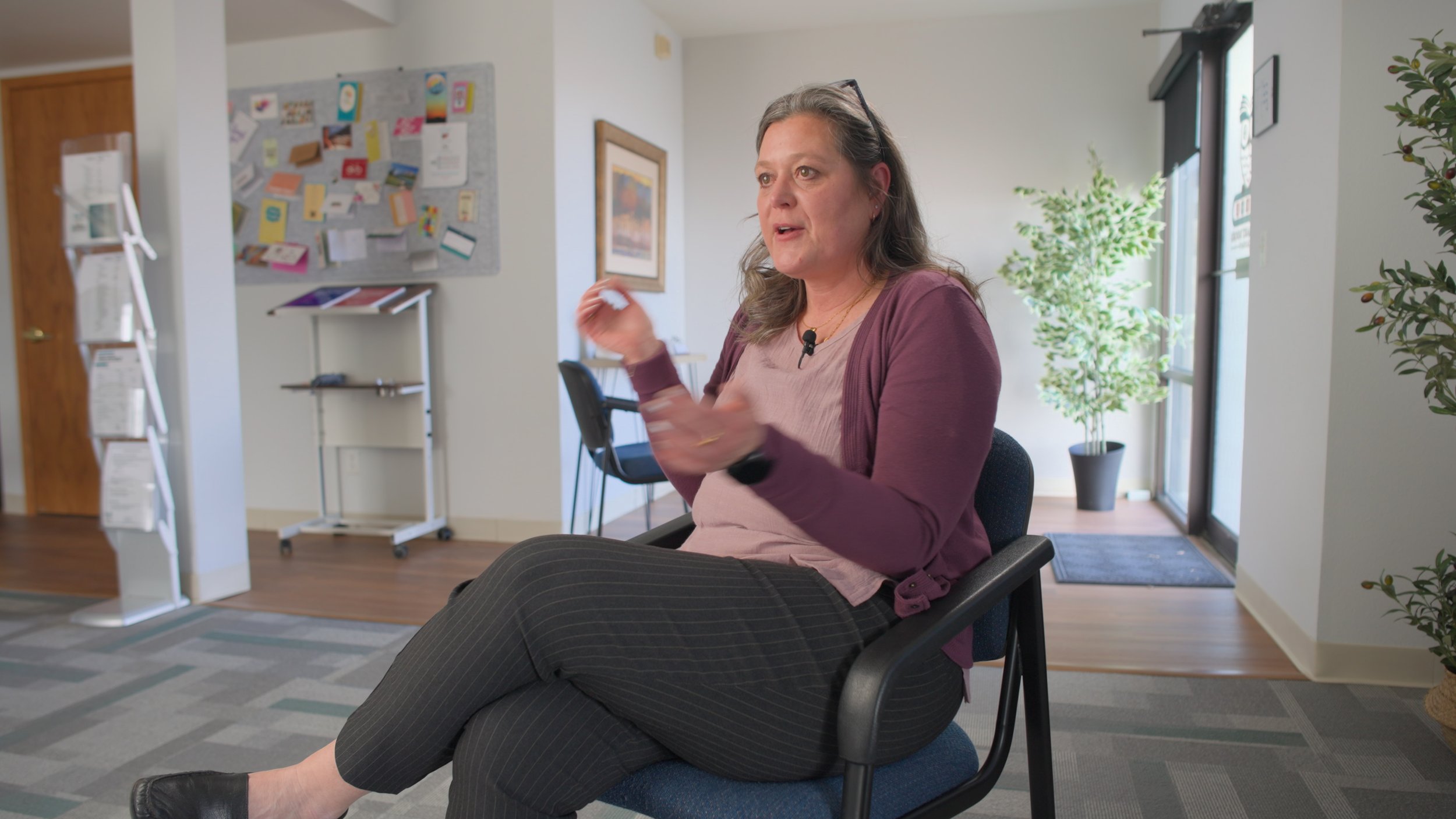 A woman sitting on a black chair in an office space, gesturing with her right hand while speaking. She has gray hair, is wearing glasses, a mauve cardigan, a light pink top, and black pinstripe pants. There is a bulletin board with colorful notes and images on the wall behind her, and a window with natural light to her right.