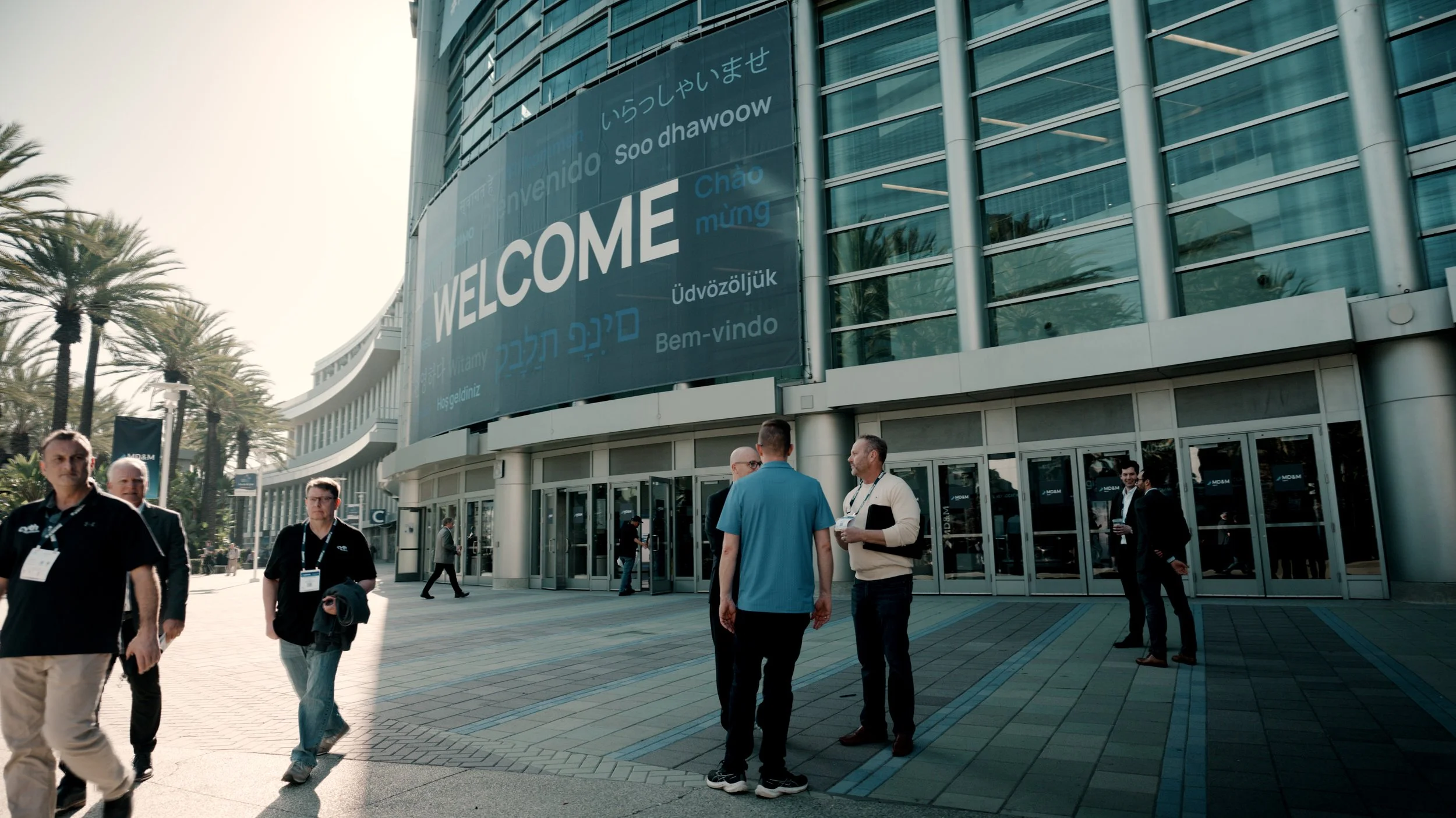 People walking outside a modern building with large glass windows and an electronic welcome sign displaying greetings in multiple languages.