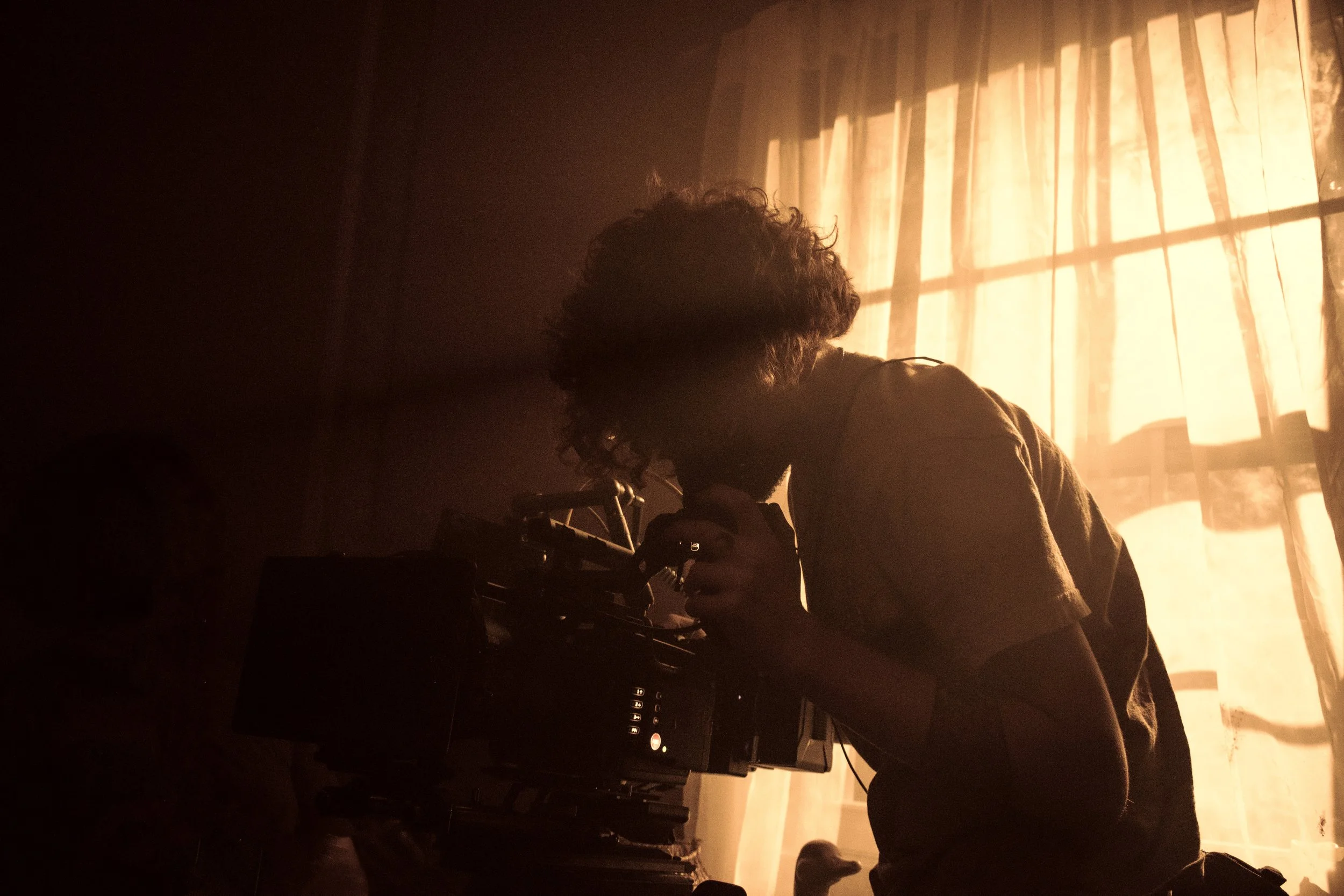 Person with curly hair sitting at a DJ setup in a dimly lit room near a window with curtains, illuminated by warm sunlight.
