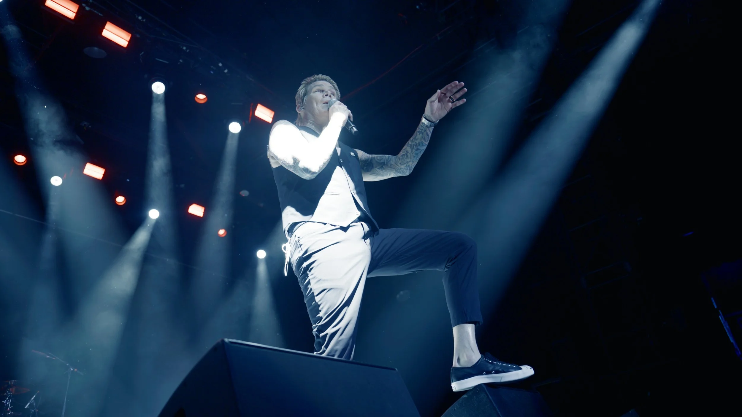 Performer on stage singing into a microphone, one foot on a monitor, illuminated by stage lights with smoke effects.