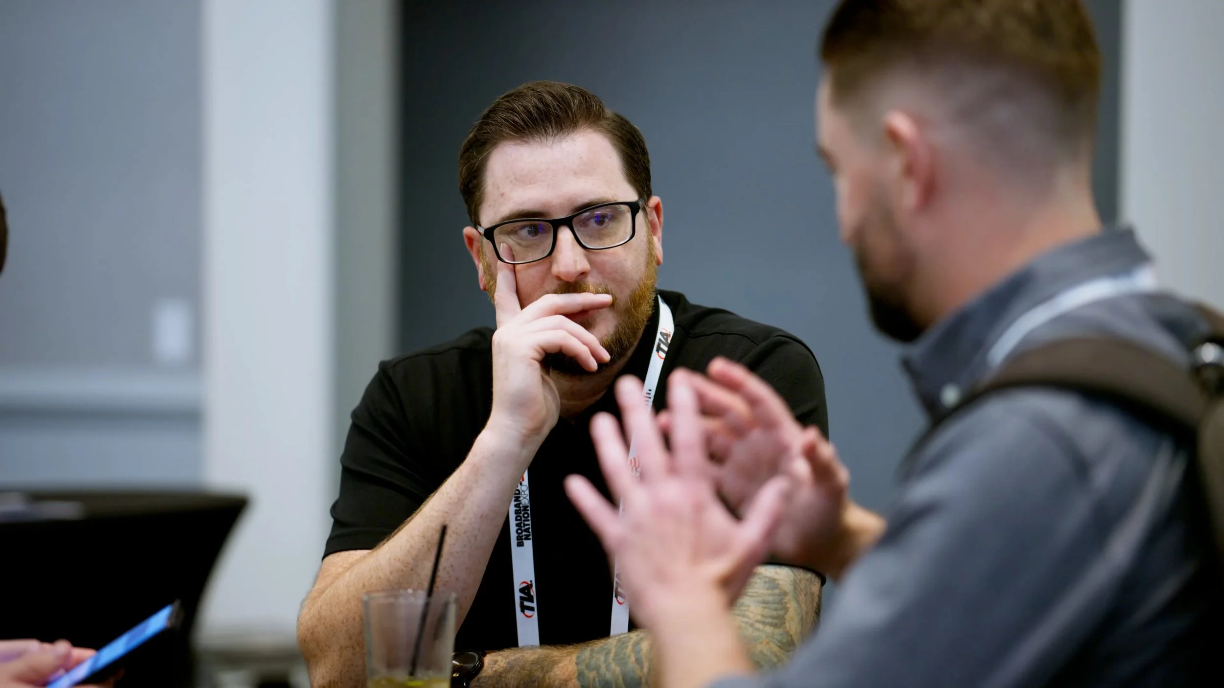 Two men having a conversation at a table. One of them is wearing glasses and appears to be listening thoughtfully, while the other is gesturing with his hands.