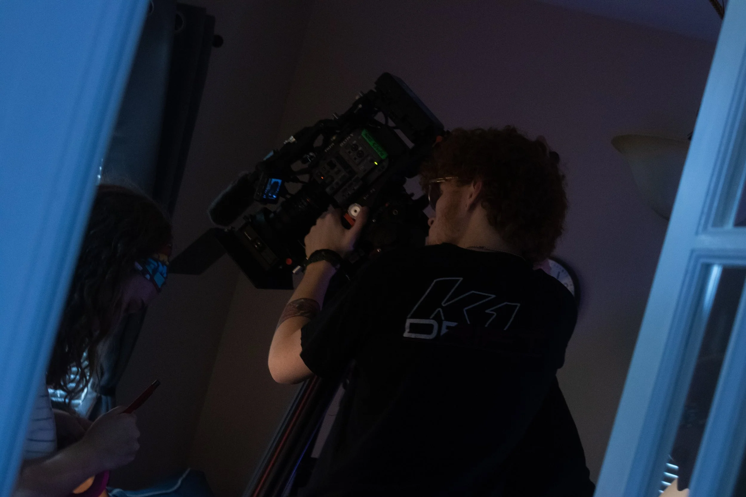 A person with curly hair and glasses operating a professional video camera in a dark room.