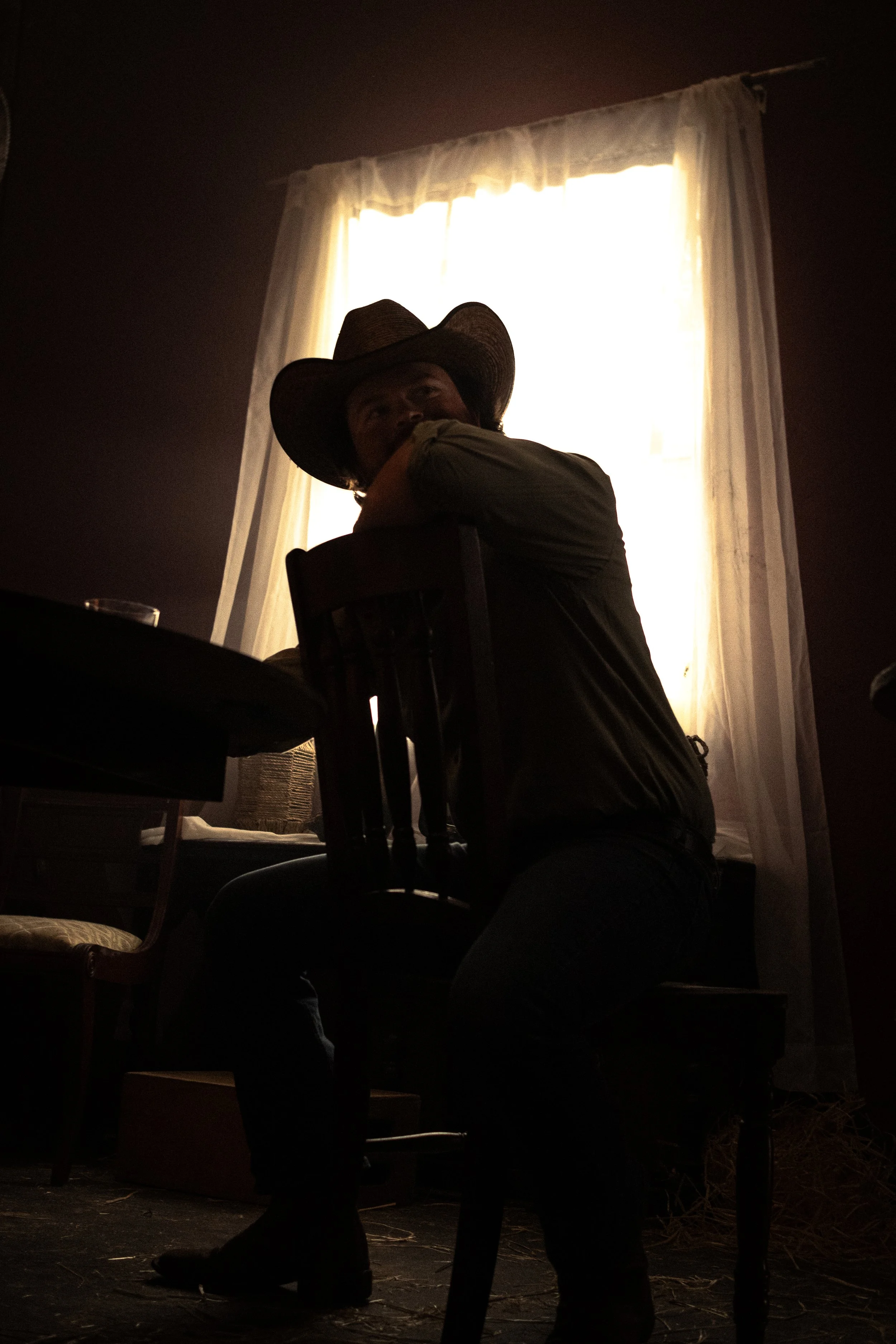Person wearing a cowboy hat sitting at a table inside a room with a window covered by sheer curtains, bright light coming through the window.