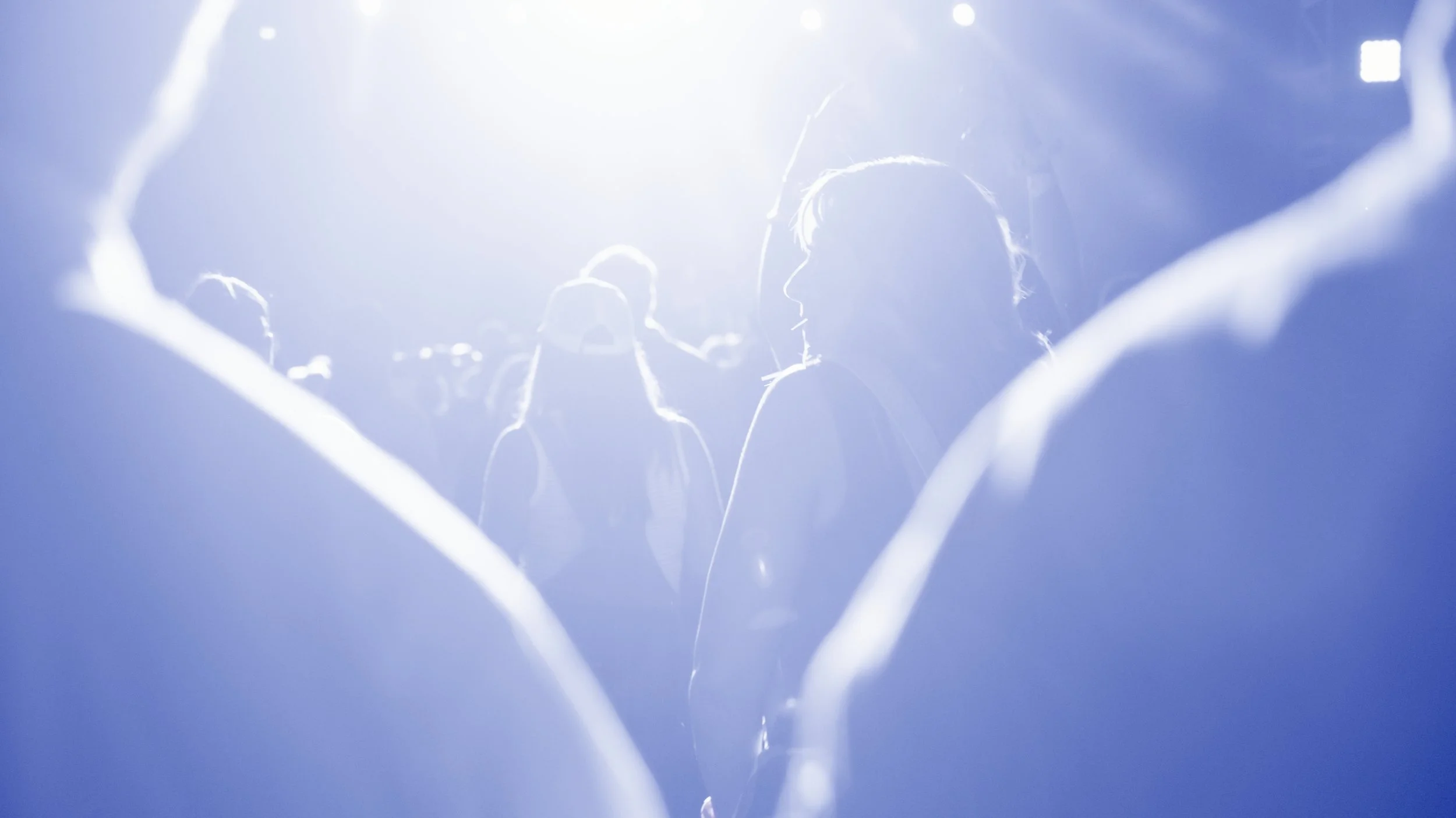 People are waiting in line or gathered in a brightly lit area with a blue tint, possibly at an event or concert, with silhouettes and an overexposed background.